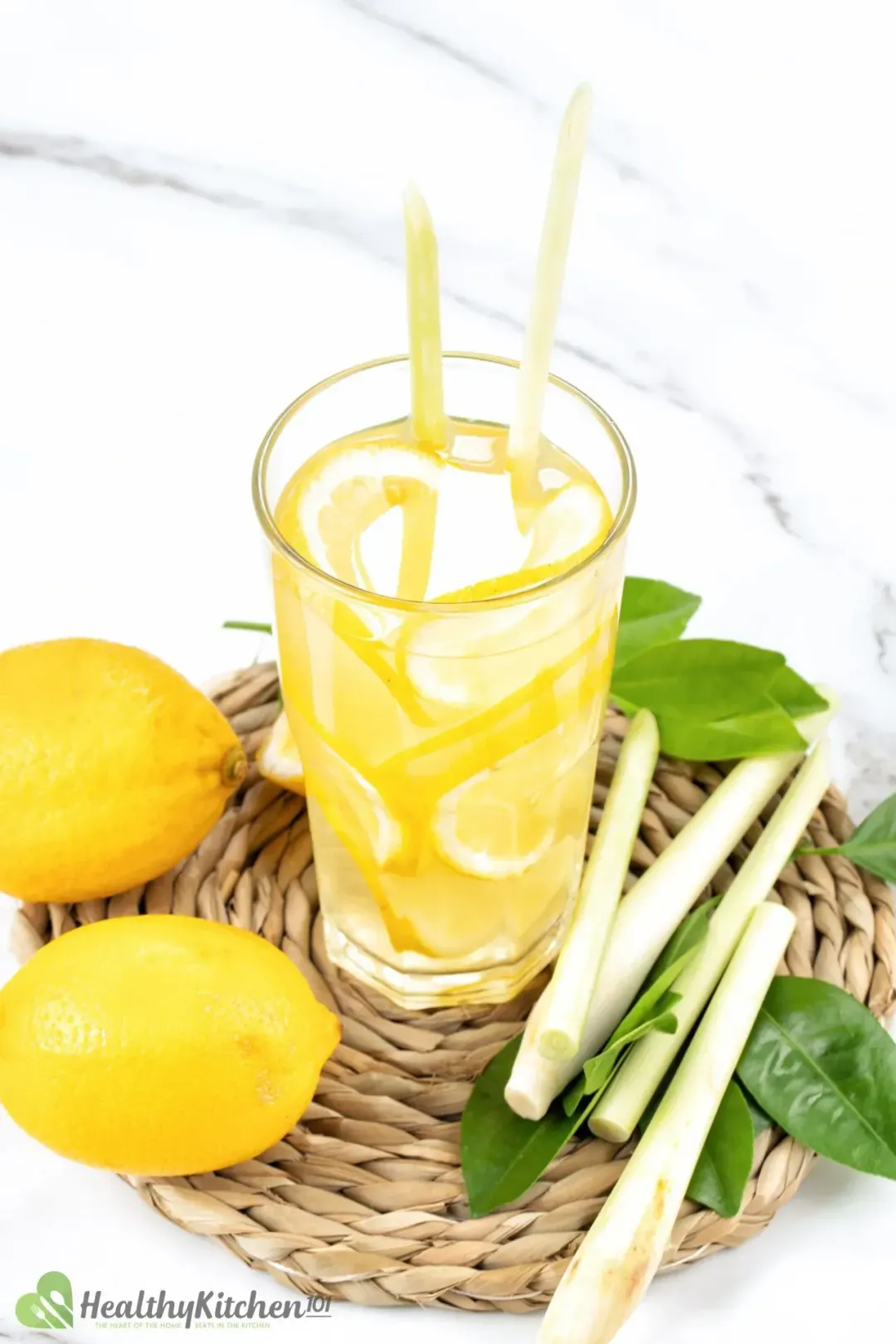 A tall glass of apple cider vinegar and lemon juice drink, packed with lemon wheels, next to lemongrass, whole lemons, and leaves