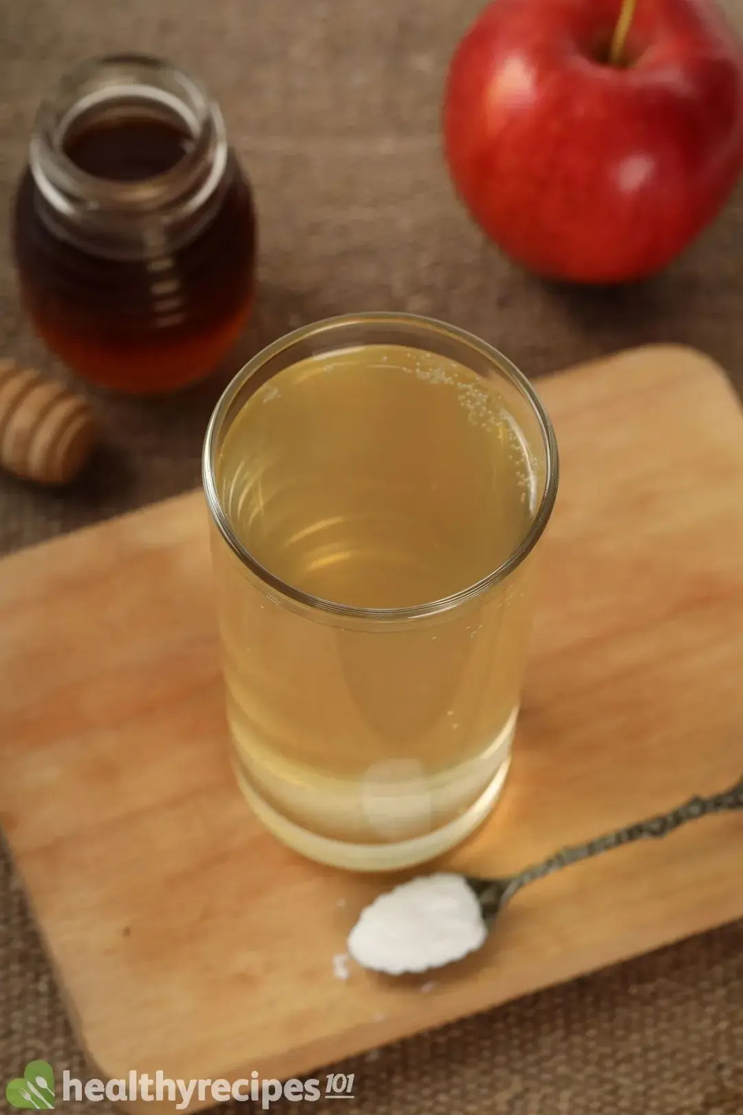A tall glass of diluted apple cider vinegar, next to a spoonful of baking soda, a honey jar, and a red apple