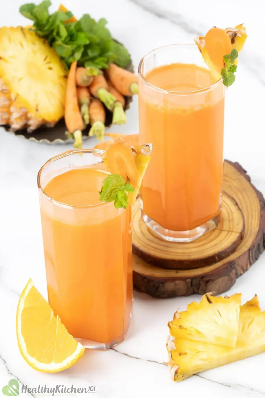 Two tall glasses of carrot orange drinks garnished with pieces of orange, pineapples, and carrots