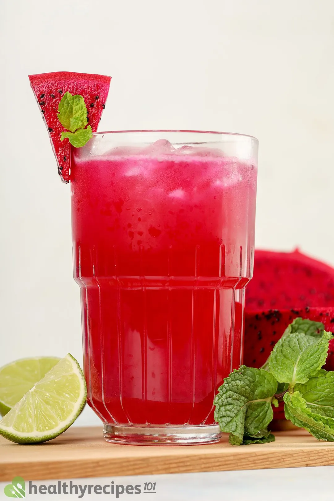 A vibrant pink glass of dragon fruit juice laid on a wooden chopping board with lime wedges, half a dragon jucie, and mint leaves.