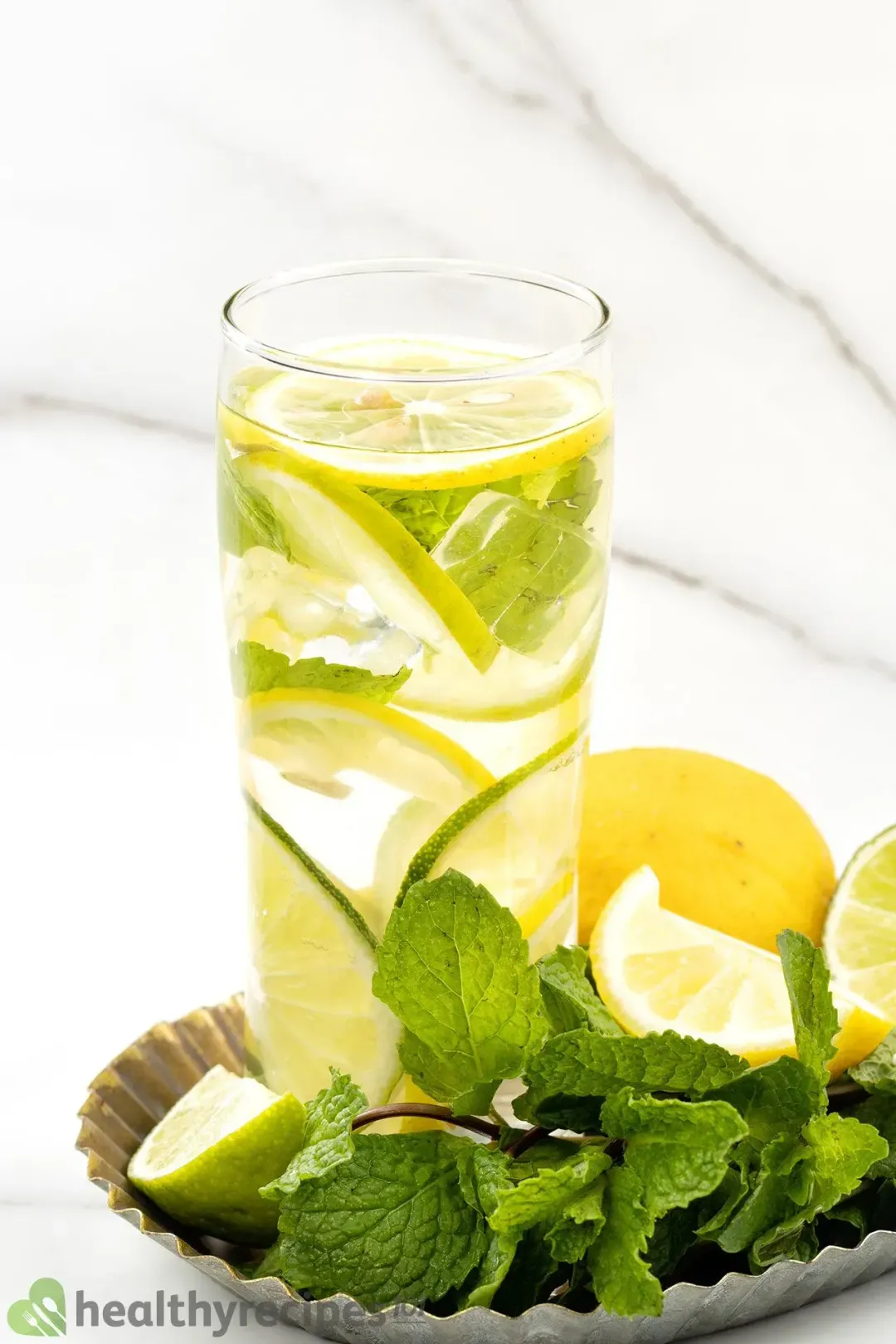 A water glass filled with lemon and lime wheels, on a metal round tray with lots of mints and citruses