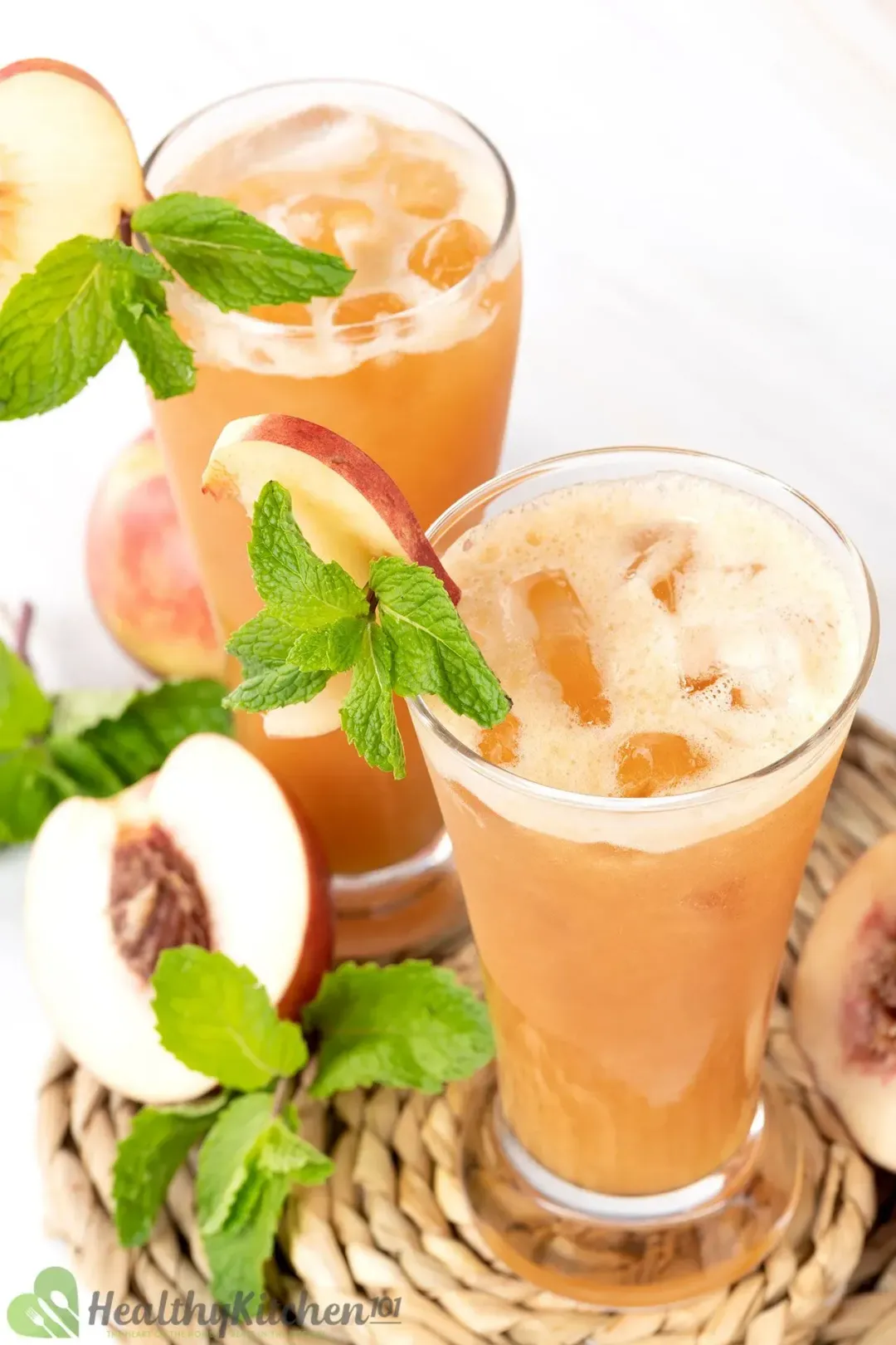 Two glasses of peach juice with ice and mint decorated on the rim, next to more mints and peach half