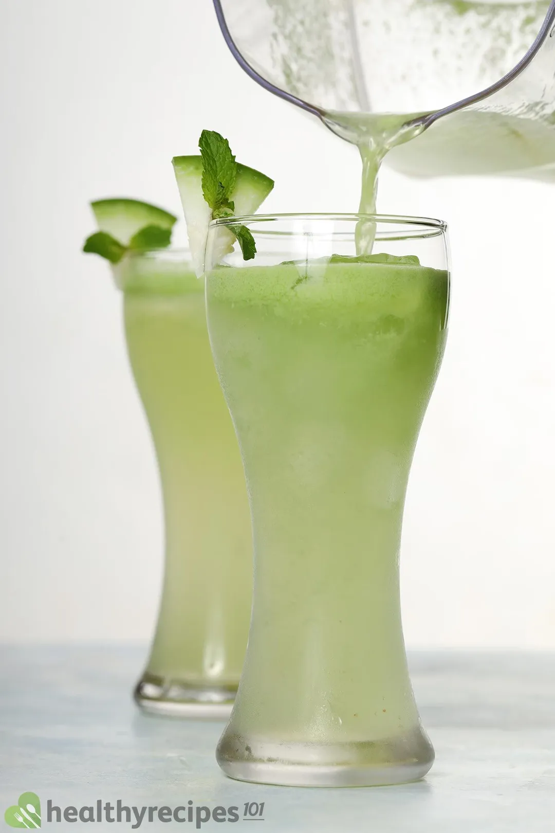 A glass jug pouring winter melon juice into a tall glass. There's a near identical glass in the back.