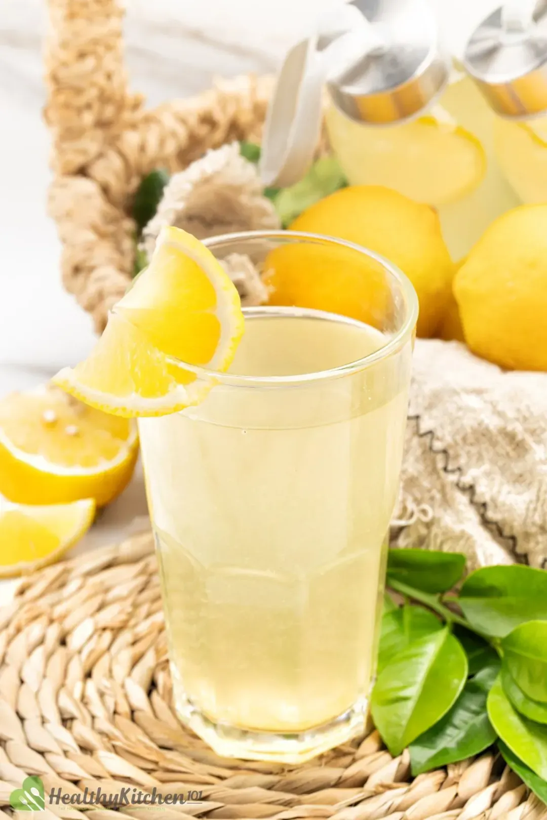 A glass of lemonade with a lime wedge on its rim, put near some lemon leaves, whole lemons, and some decorations