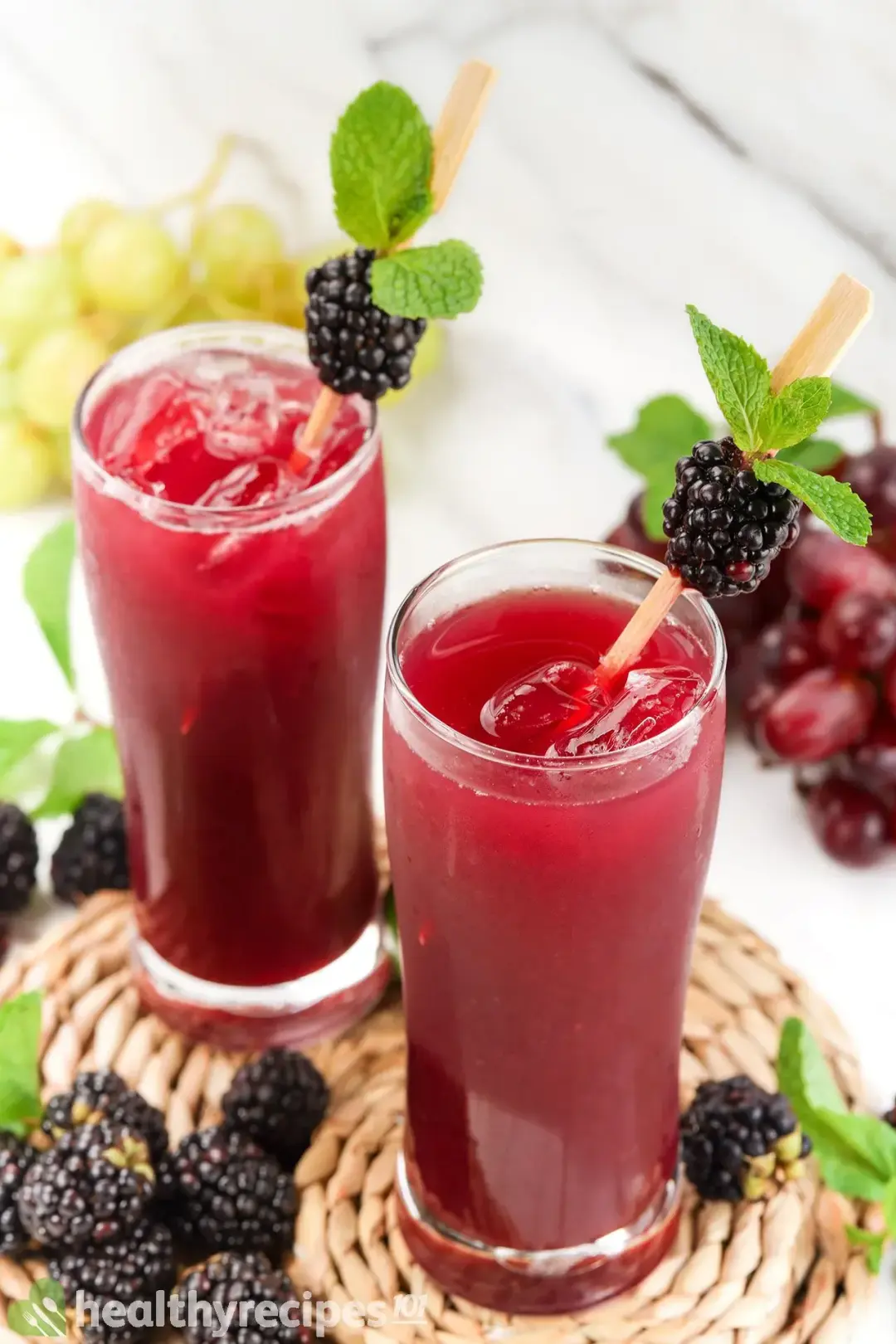 Two iced glasses of black berry juice drinks, with straws, mints, put next to a bunch of blackberry