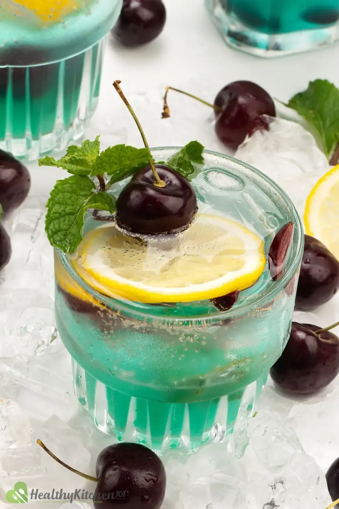 Cocktail glasses of a cyan-hued drink, topped with cherries and lemon wheels, next to scattered cherries