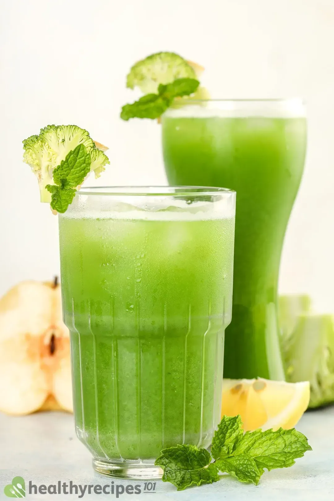 Two glasses of broccoli juice drinks garnished with lime wedges, mints, and next to some fruits