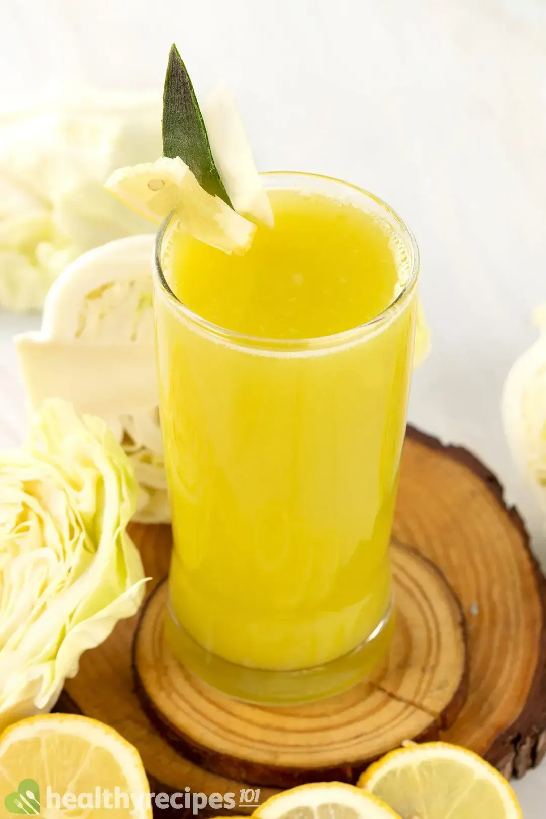 A glass of yellow-green cabbage drink, next to some cabbages and lemon wheels