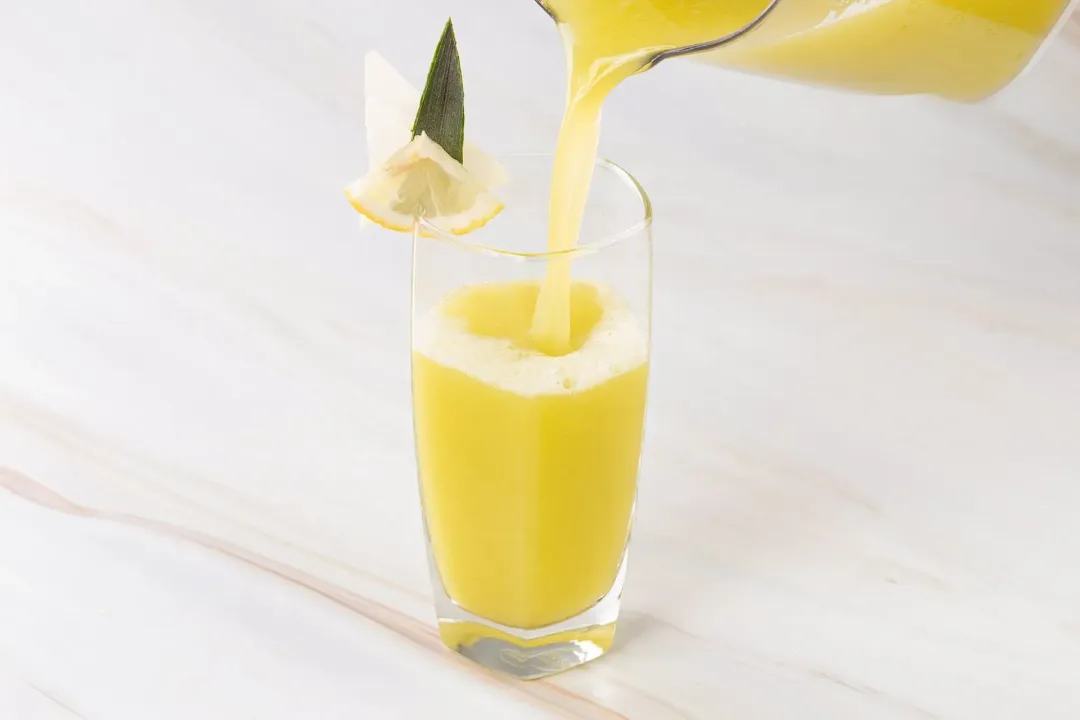 A glass of yellow-green cabbage drink poured from a pitcher above