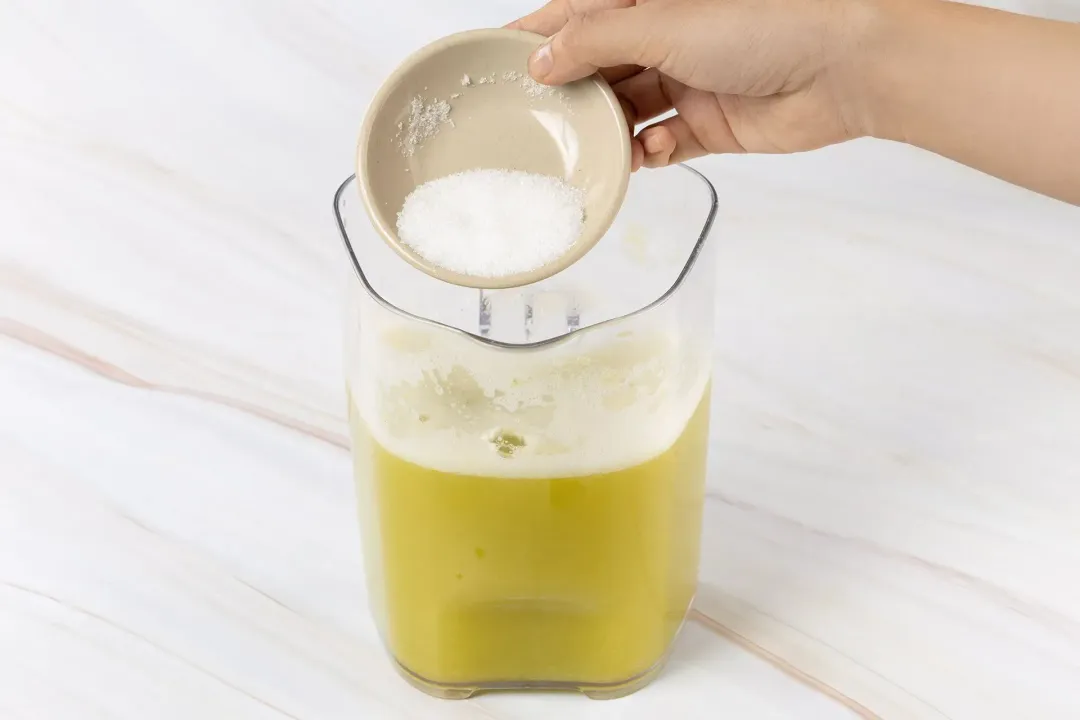 A hand pouring sugar into a pitcher filled halfway with a yellow-green juice