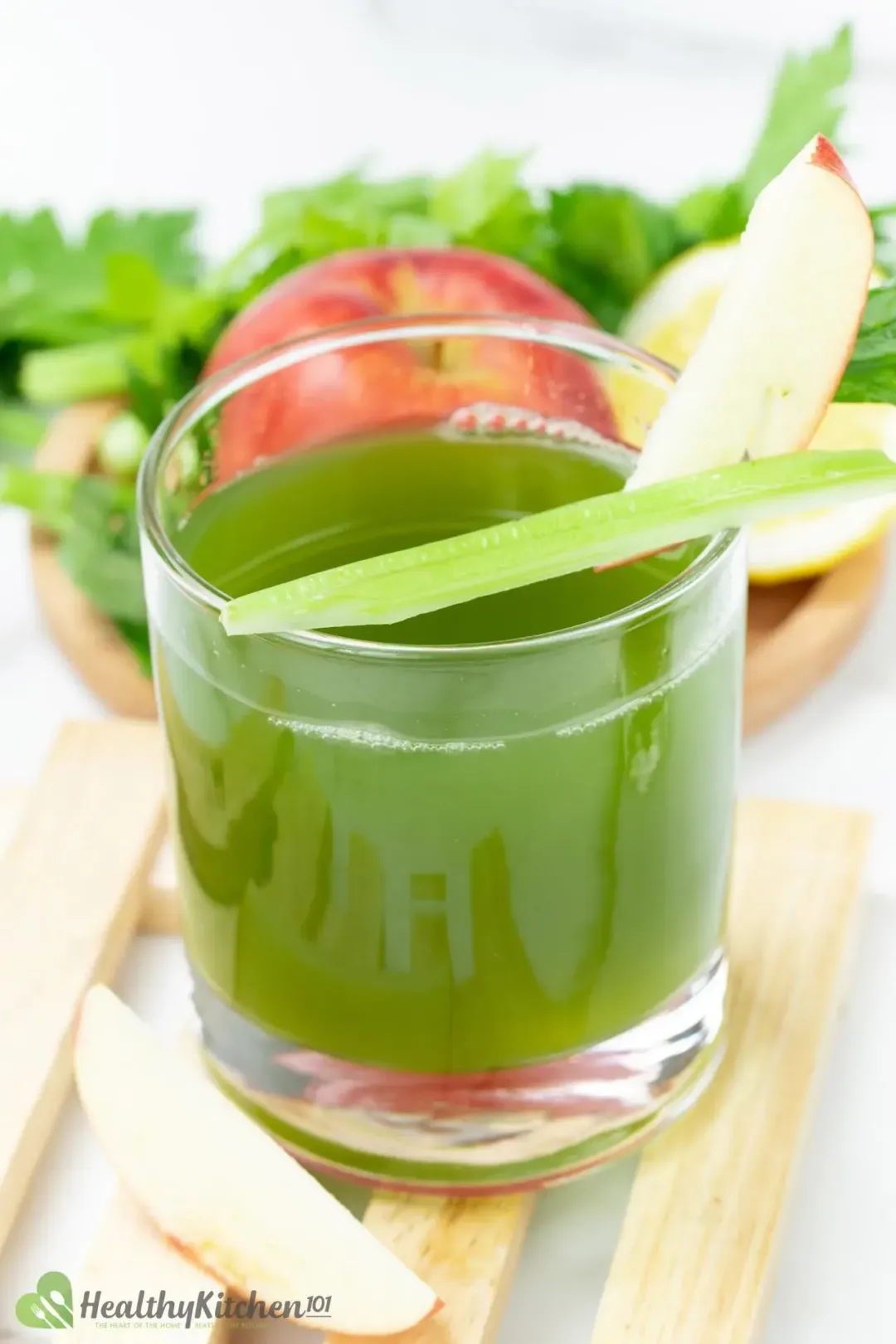 A glass of celery juice with an apple wedge and a celery stalk on the rim, garnished with celery leaves