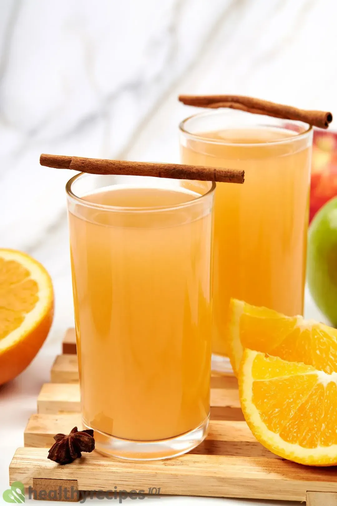 Two glasses of apple cider with cinnamon sticks on top, next to some orange wedges