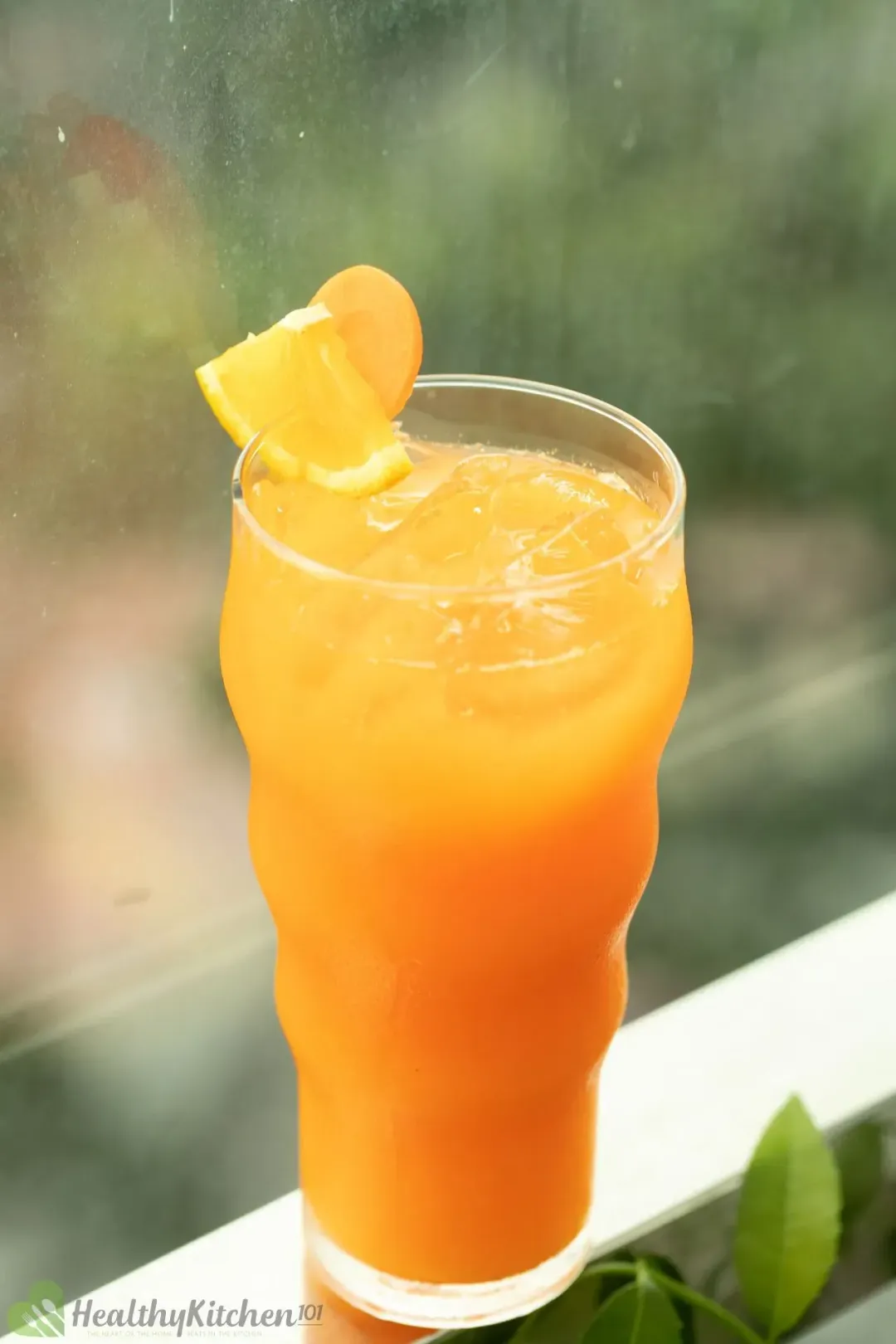 Three glasses of iced carrot orange drinks of different sizes, next to orange wedges and some baby carrots