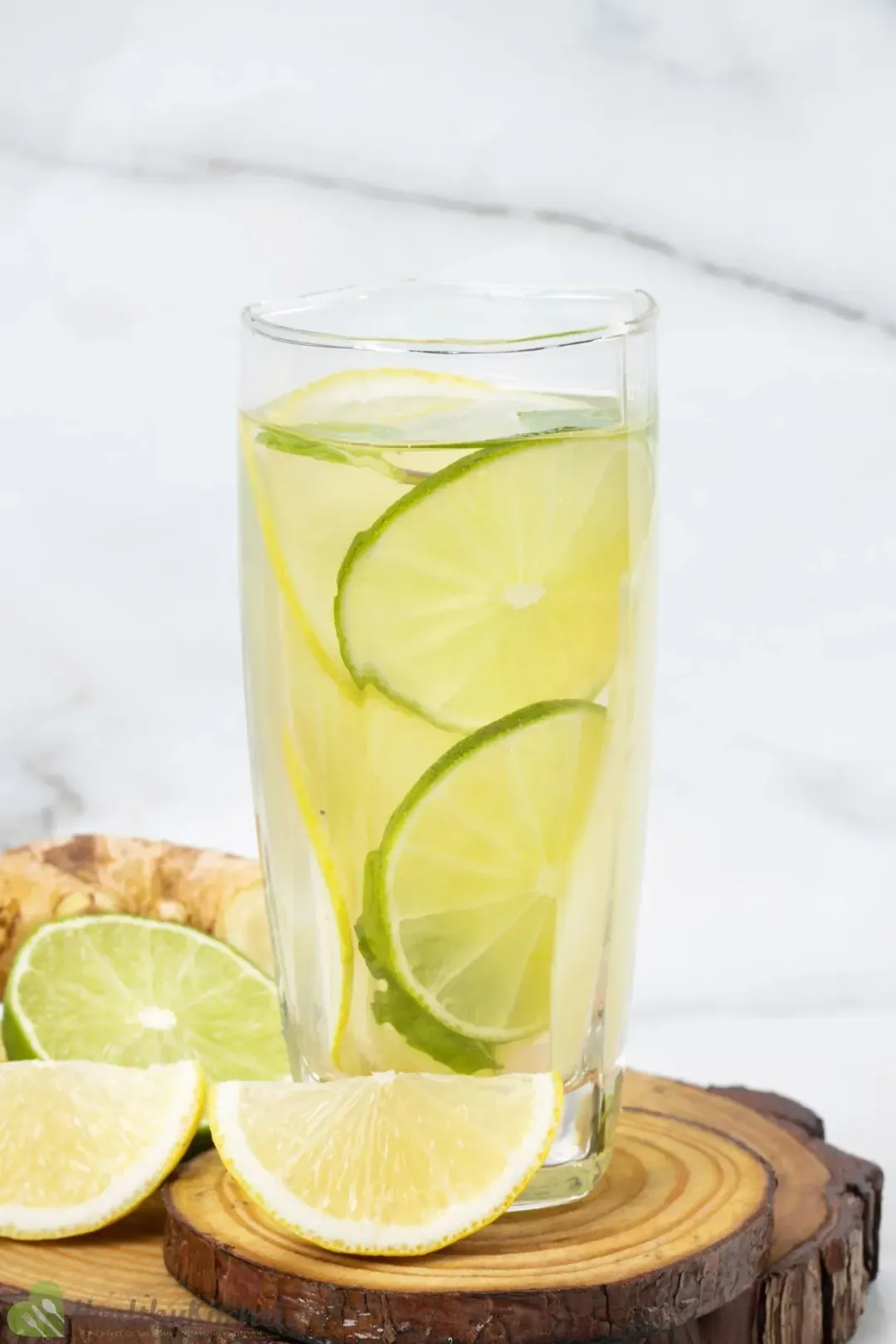 A tall glass of clear water submerging lemon and lime wheels, put on wooden coasters next to lemons and limes
