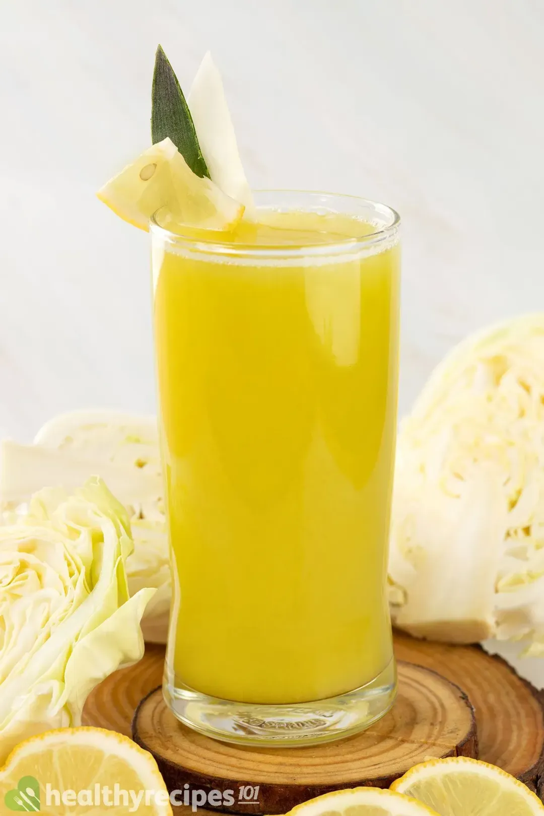 A glass of yellow-green cabbage drink, next to quartered cabbages and lemon wheels underneath