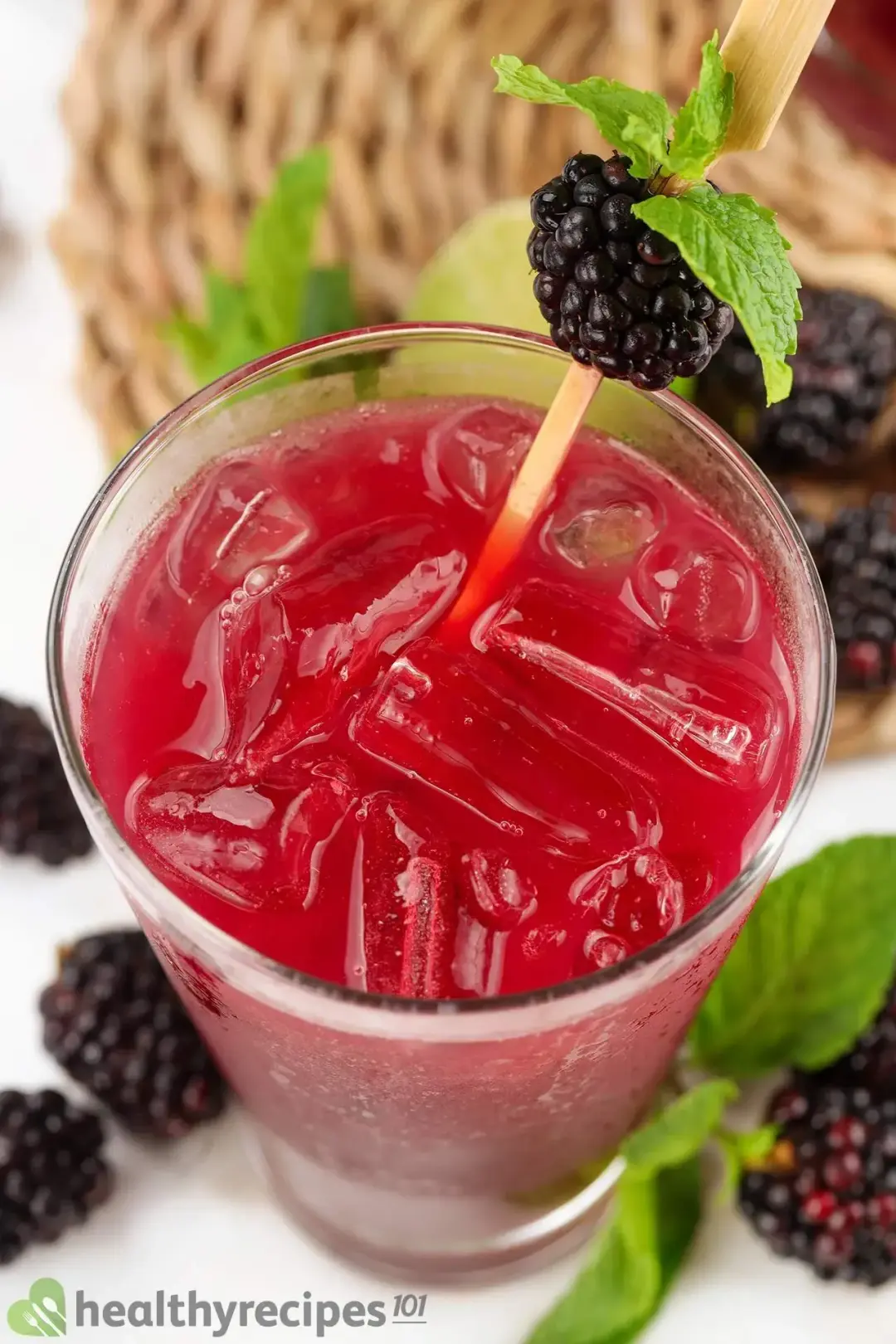 A close-up shot of a glass of red-pink blackberry drink, garnished with mints and blackberries lying around