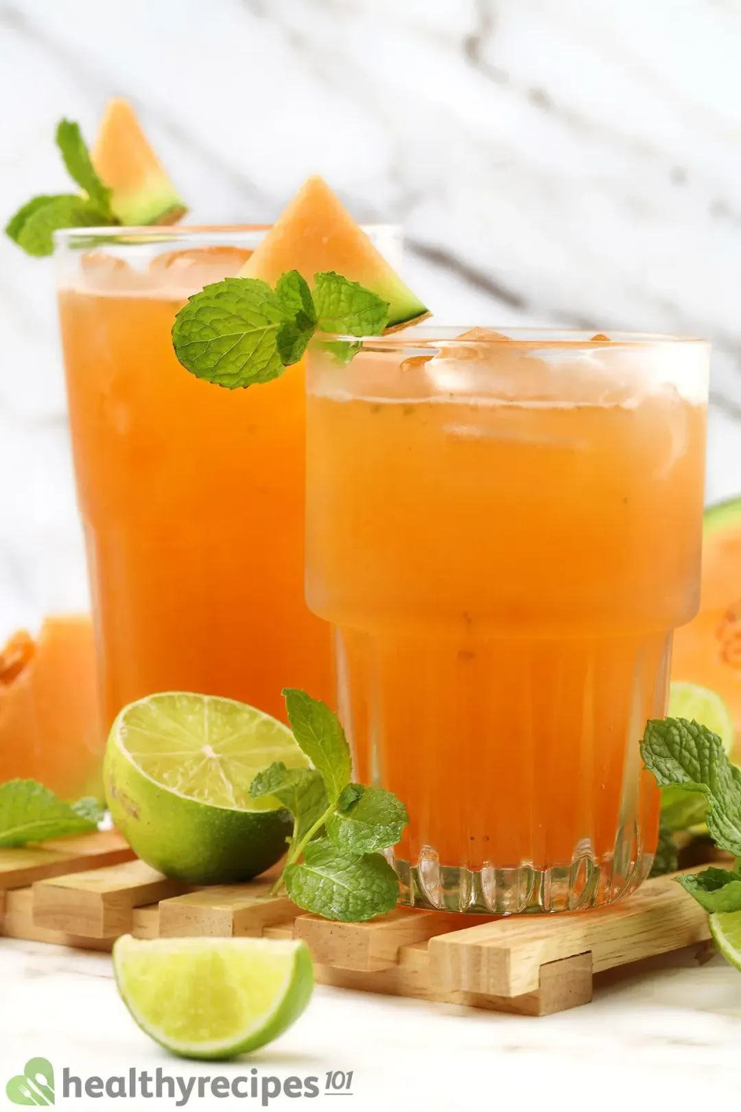 Two iced glasses of cantaloupe drinks, garnished with mints, cantaloupe wedges, and lime wedges