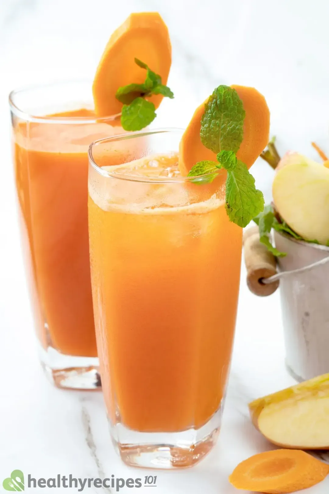 Two tall glasses of an Orange carrot drink Garnished with mint leaves, carrot slices and Apple quarters
