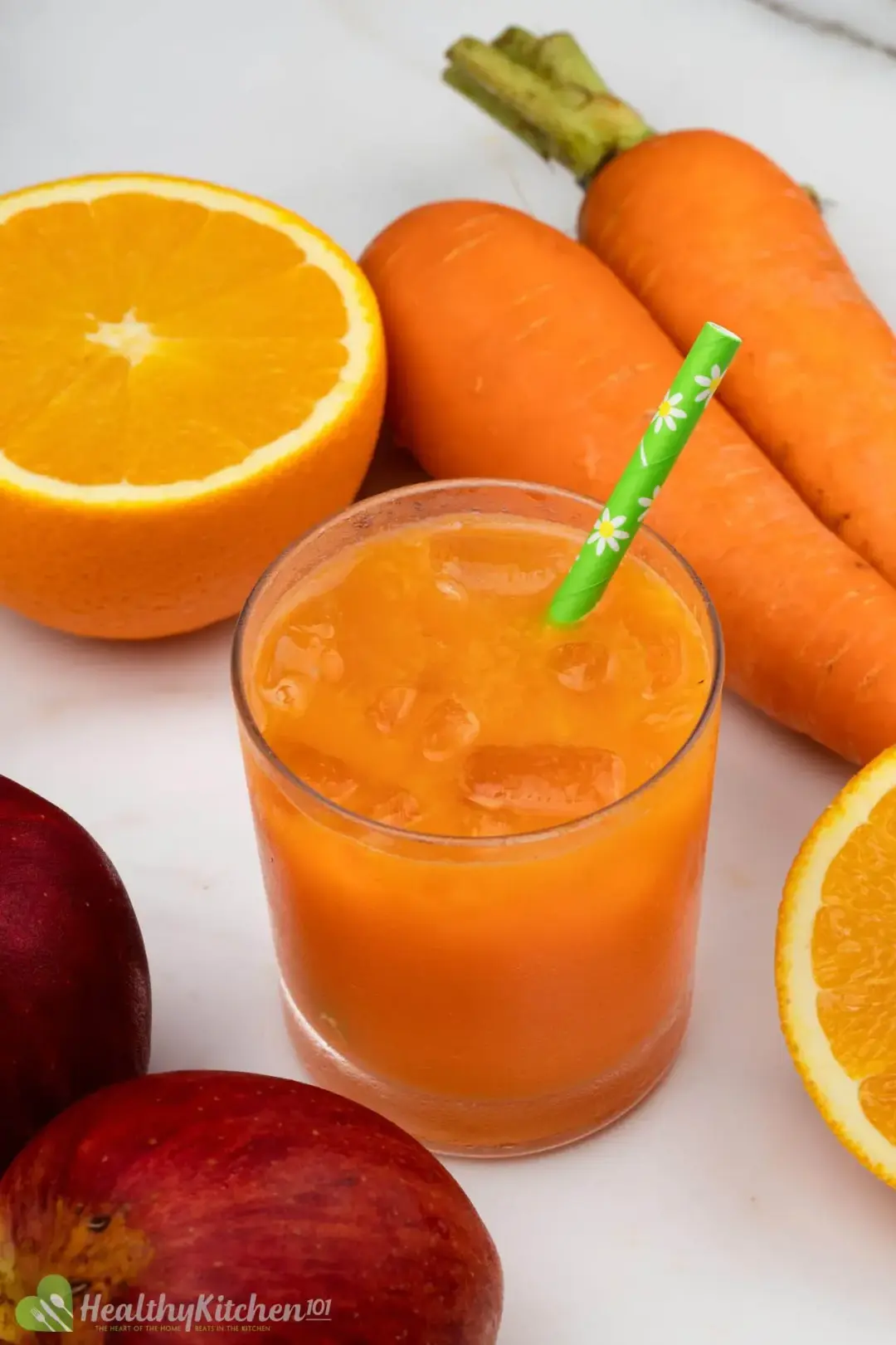 A short glass of iced carrot apple drink next to some red apples, orange halves, and whole carrots