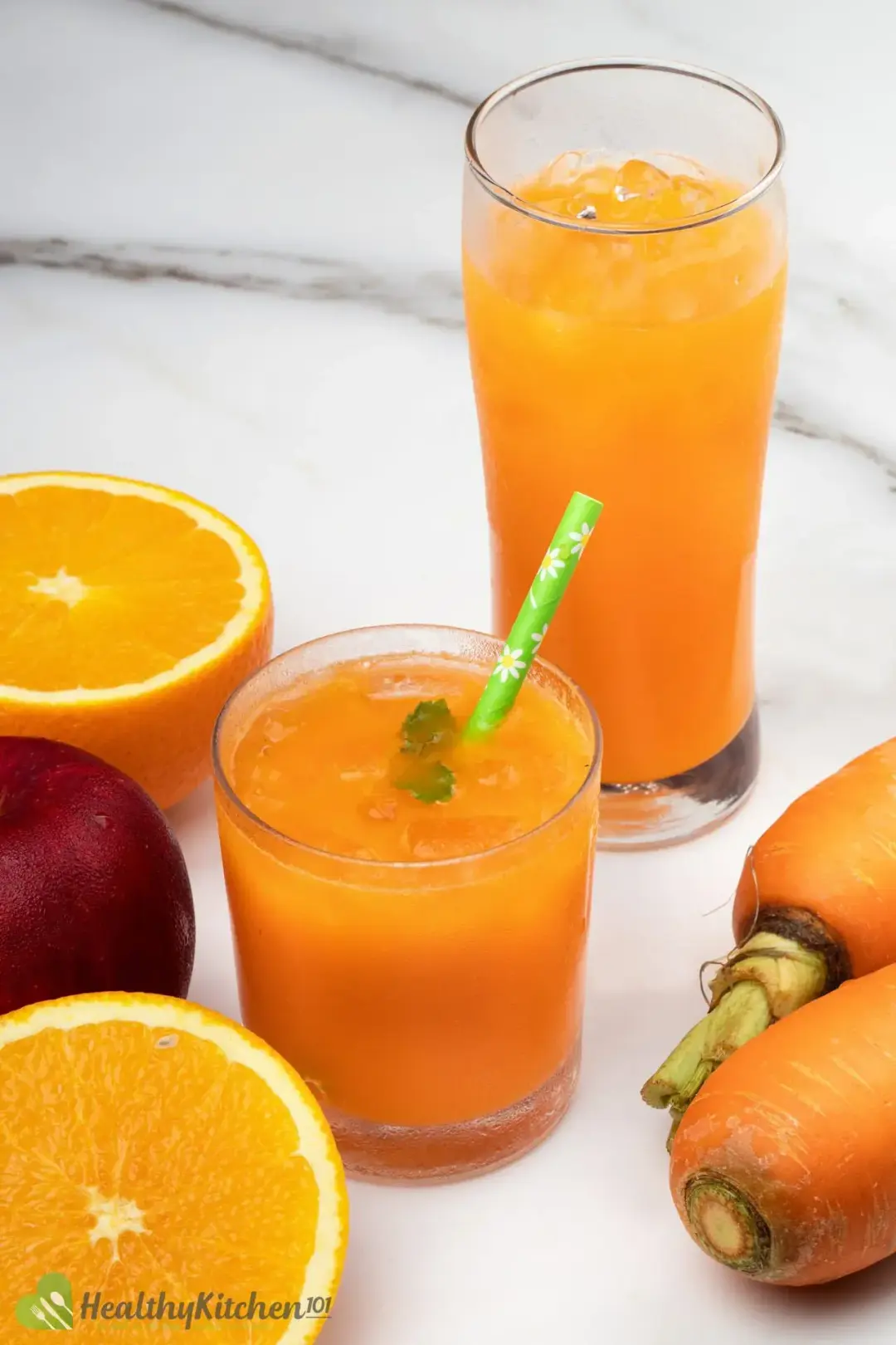 A short glass of iced orange carrot juice next to a tall glass of carrot drink, a red apple, two orange halves, and carrots
