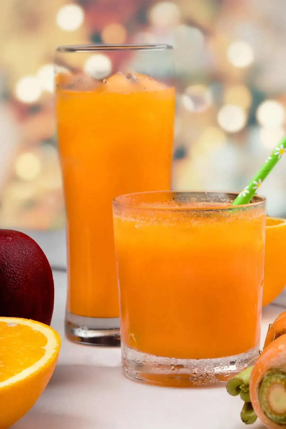 Two glasses of iced carrot apple drink, one tall and one short, next to an apple and half an orange