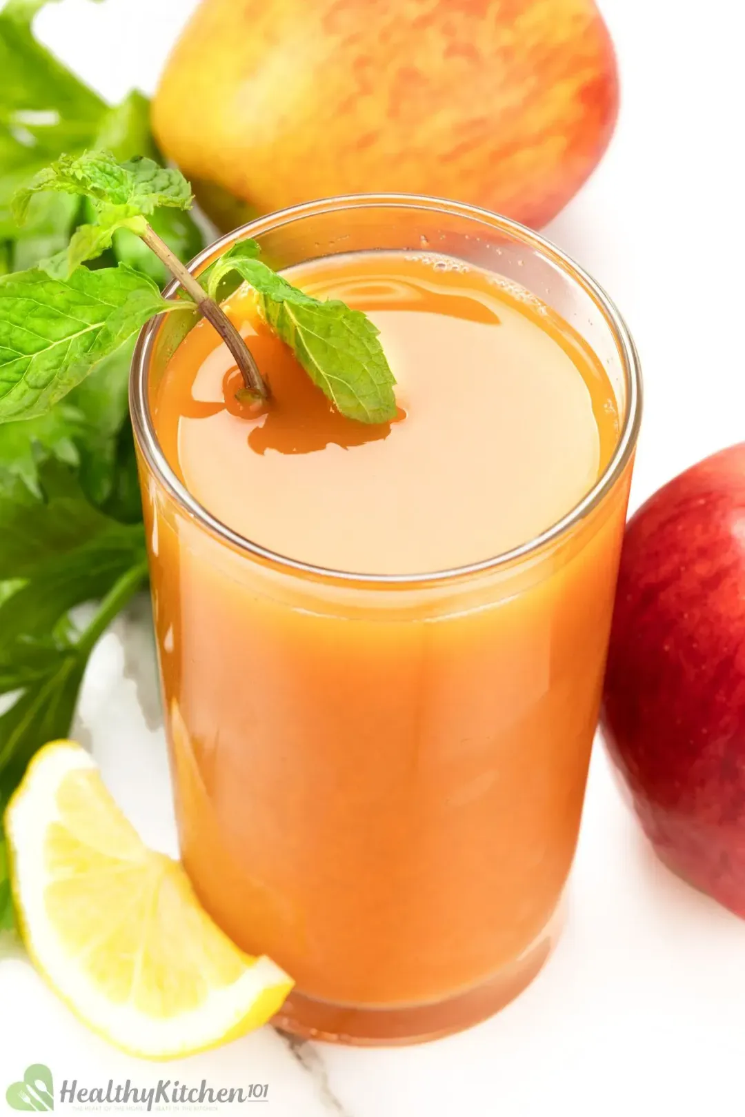 A glass of carrot juice with a mint sprig, a lemon wheel, and some apples in the back