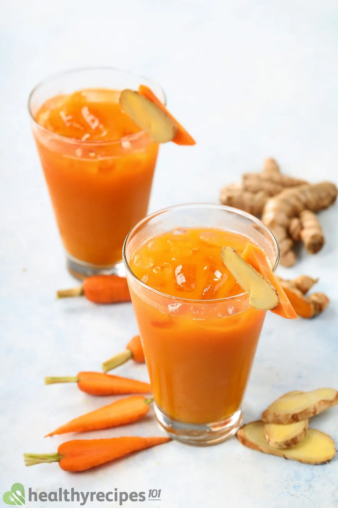Two glasses of Carrot Ginger Turmeric Juice surrounded by slices of carrots and ginger.