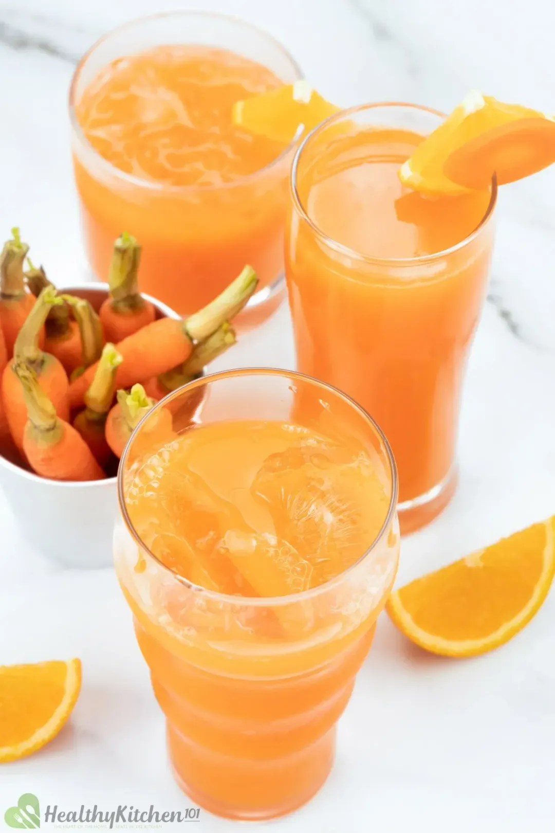 Three glasses of iced carrot orange drinks of different sizes, next to orange wedges and some baby carrots