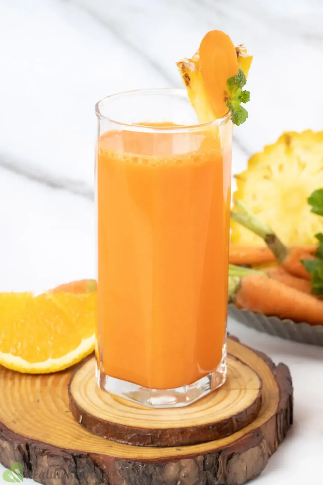 A tall glass of carrot orange pineapple juice put on wooden coasters, next to an orange wedge and some carrots