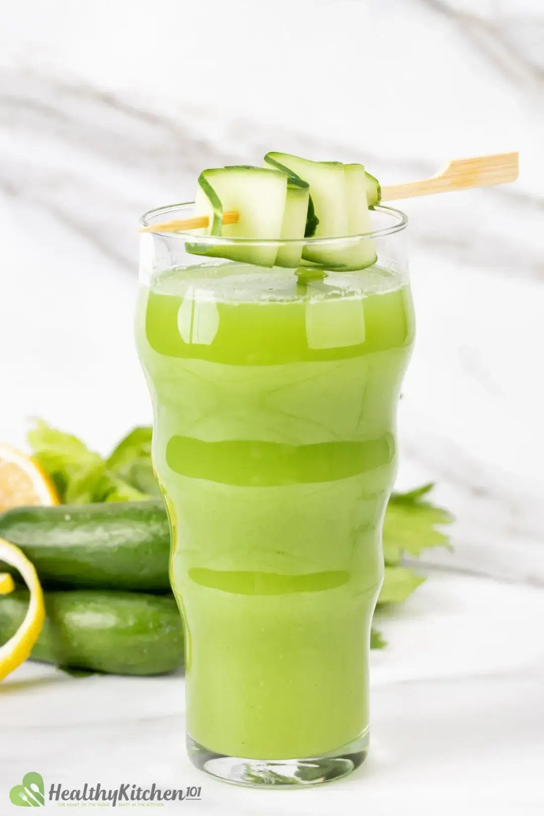 A tall glass of cucumber celery drink topped with skewered cucumber and with whole cucumbers in the back