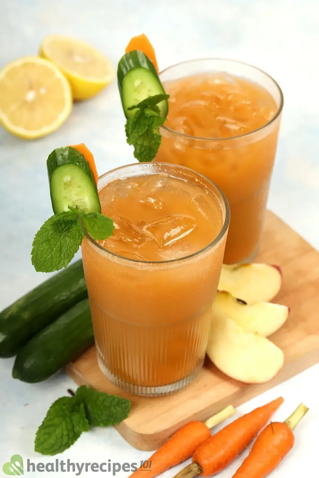 Brown drinks with ice inside two glasses garnished with cucumber slices, mint leaves, apple slices, and baby carrots