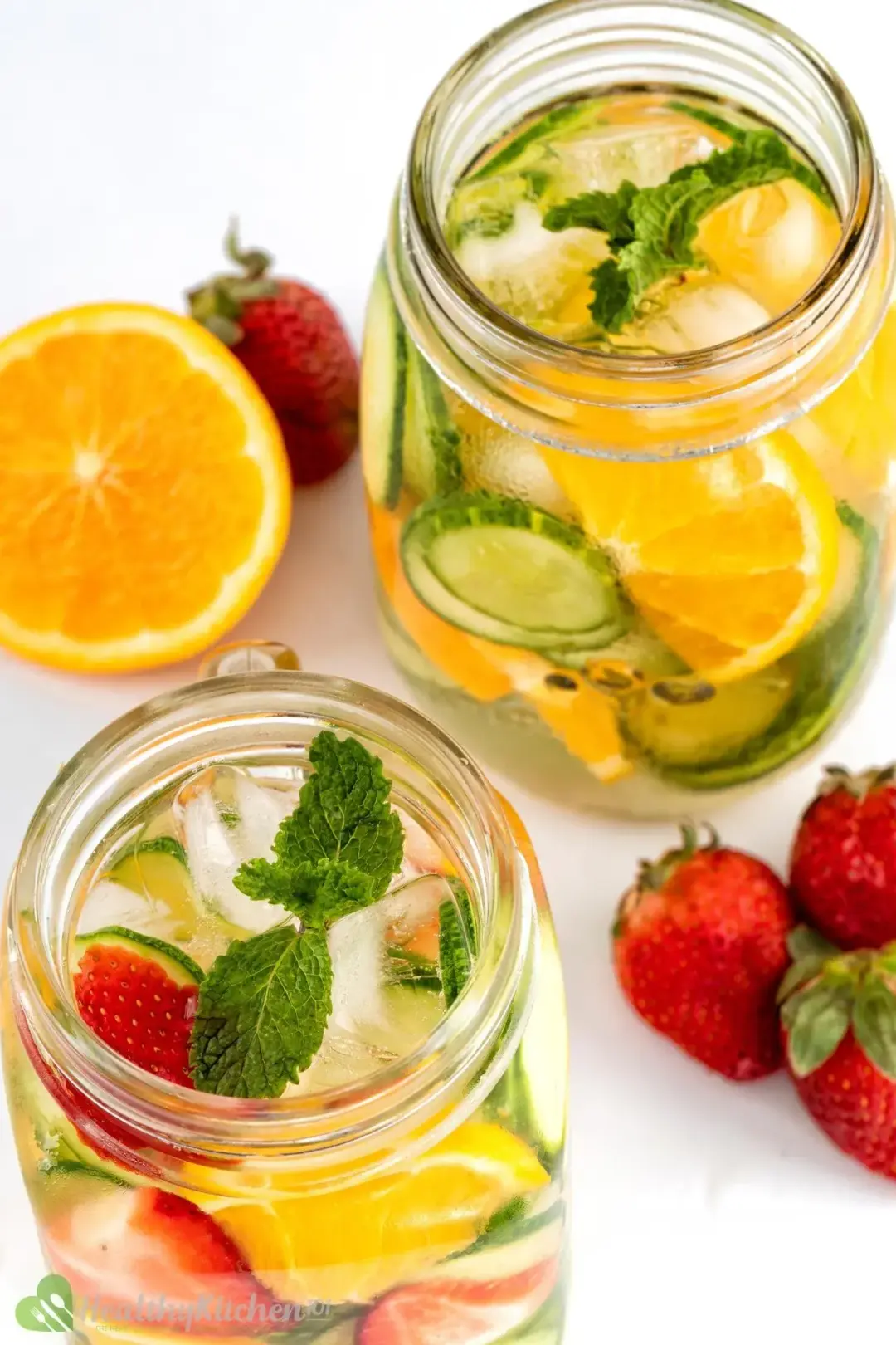 Two jars of clear water submerging strawberries, orange, mint leaves with ice