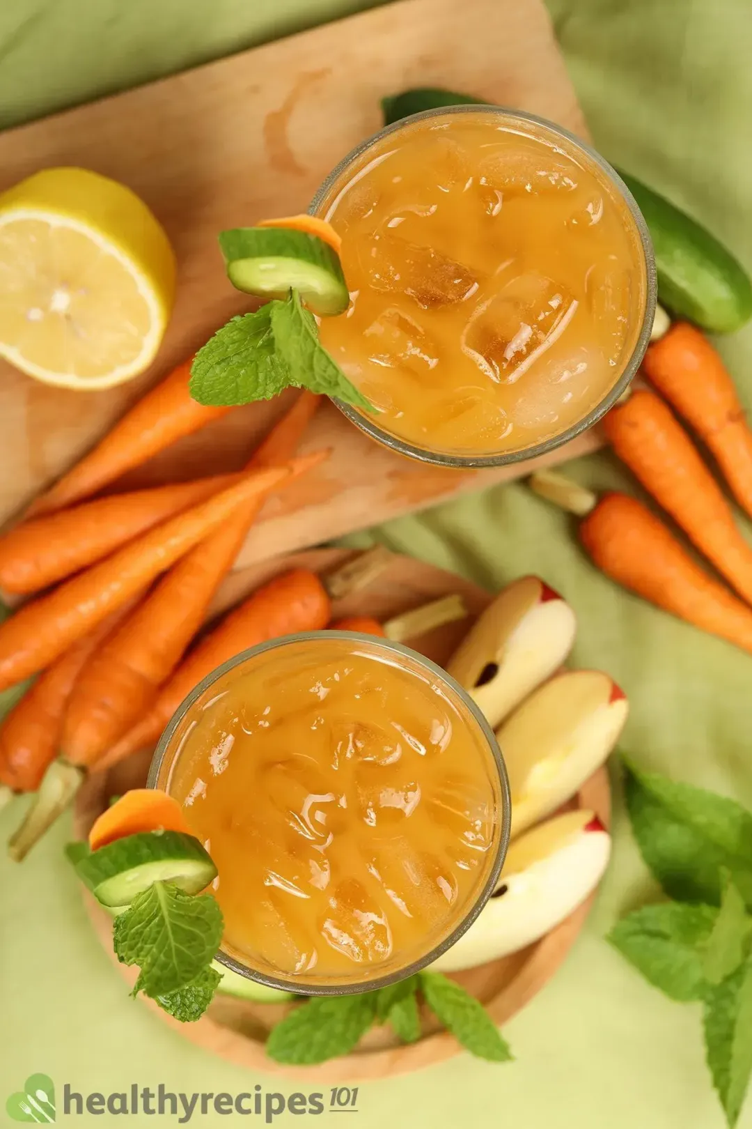 A picture from the top of two iced carrot drinks next to lots of whole baby carrots, mints, sliced cucumbers, and apple wedges