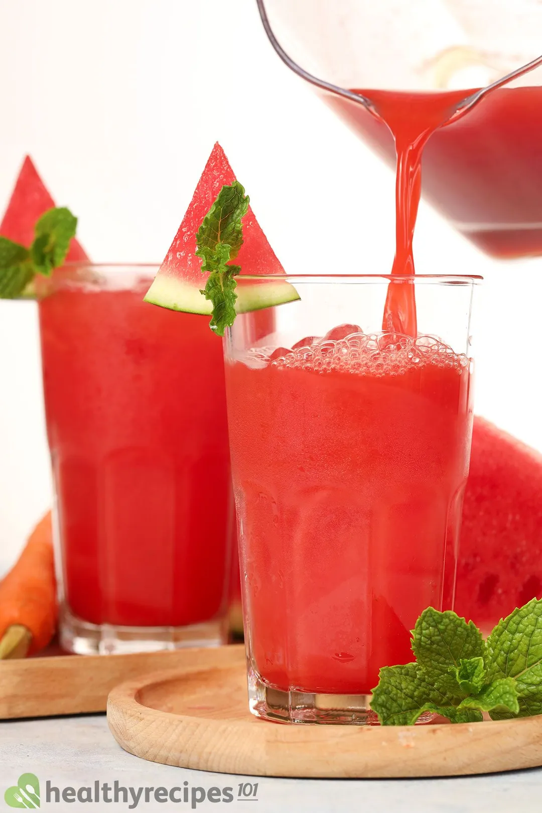 A jug pouring watermelon carrot juice into a glass placed on a wooden board with mint leaves.