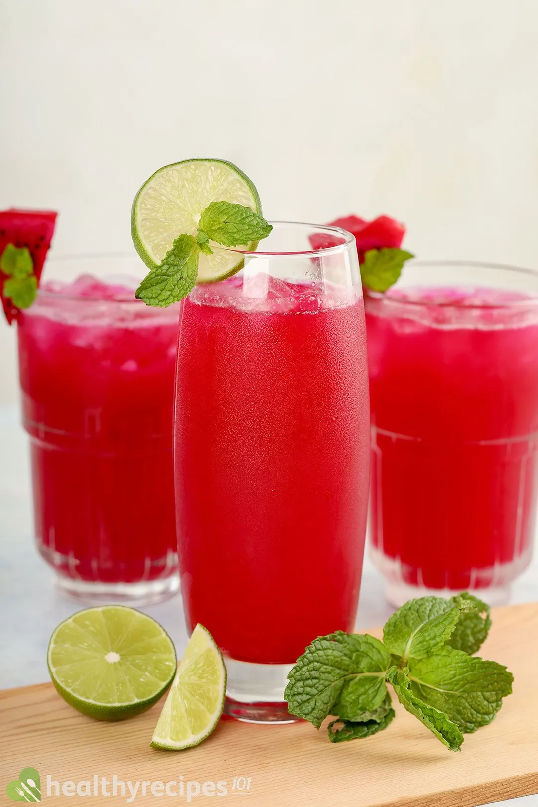 Three glasses of dragon fruit juice placed on a wooden board with lime wedges and mint leaves.