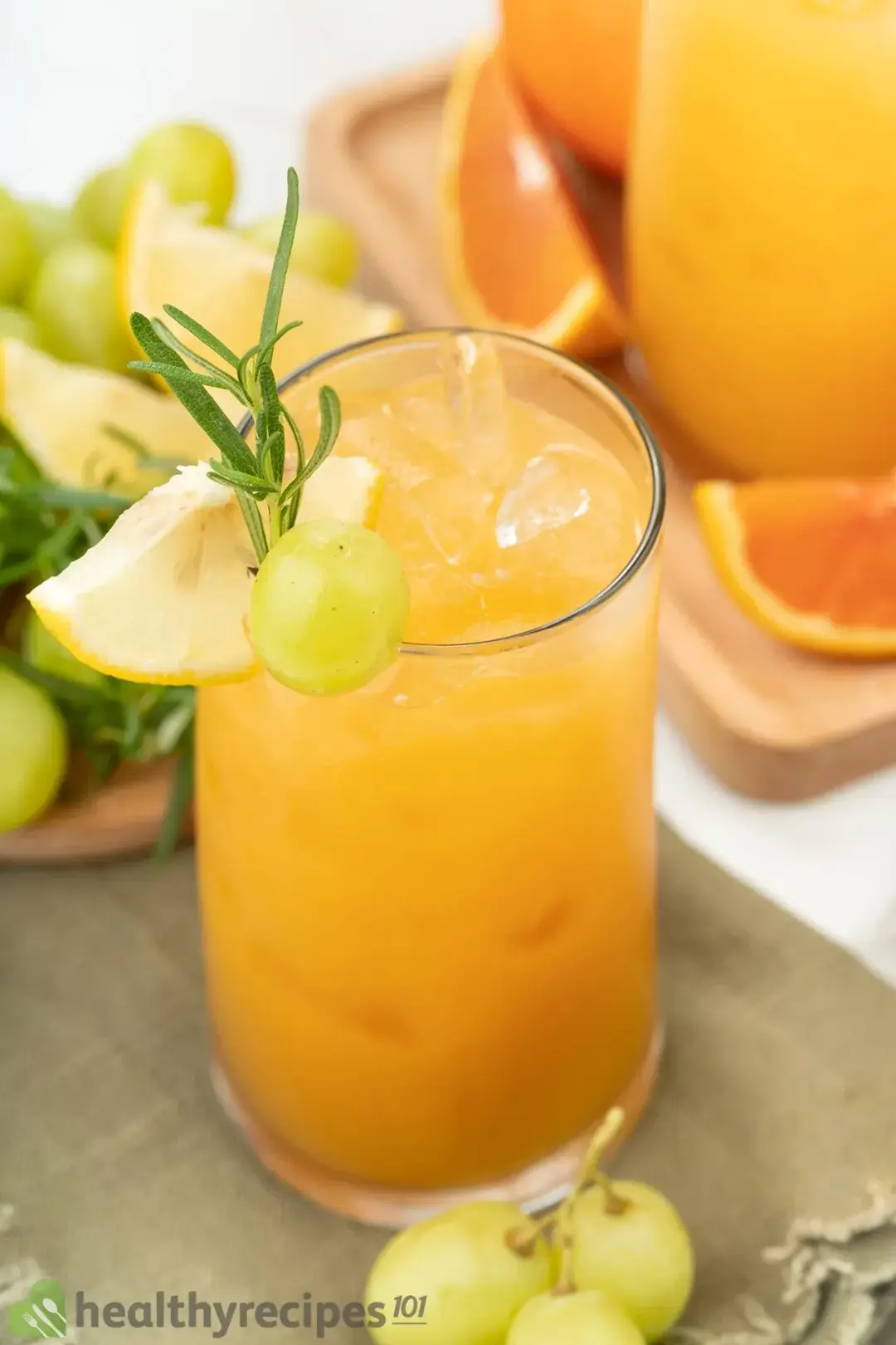 A tall glass of iced grapefruit drink topped with a rosemary sprig and put next to grapes