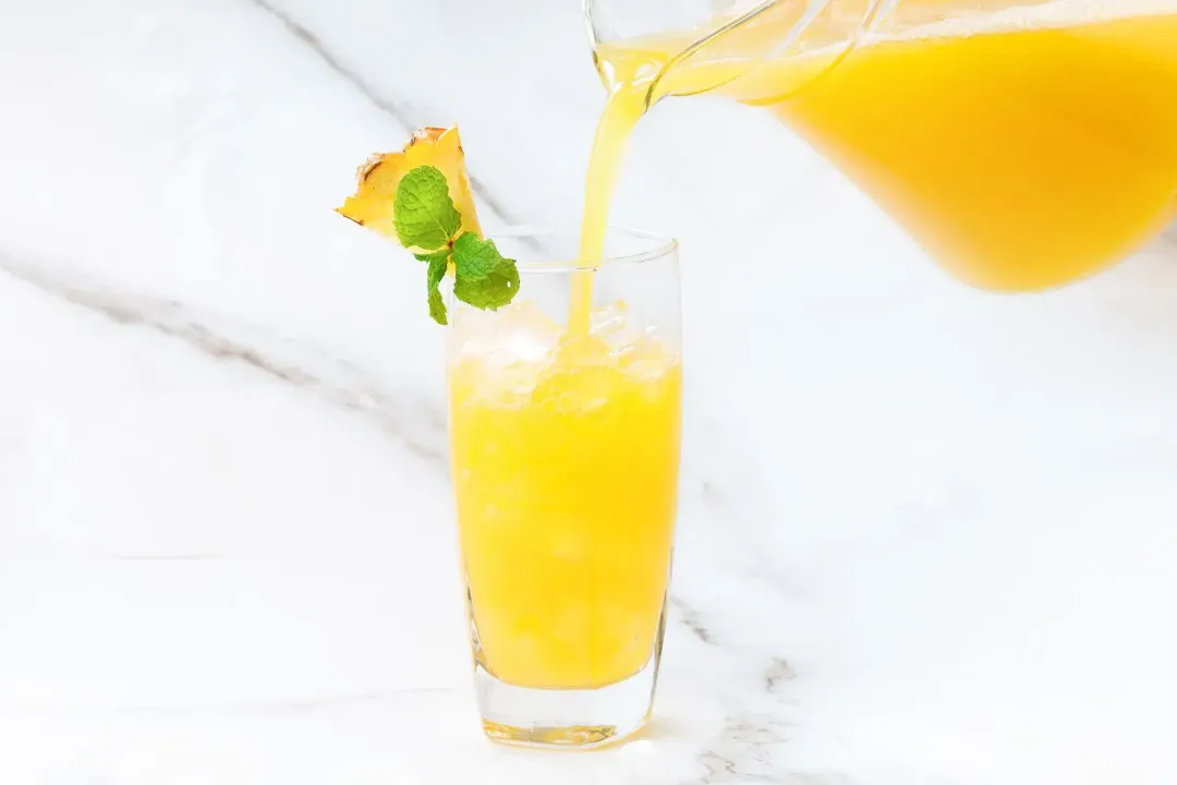 A glass pitcher pouring an orange pineapple juice and vinegar cocktail into a clear glass, garnished with mints and pineapple wedge