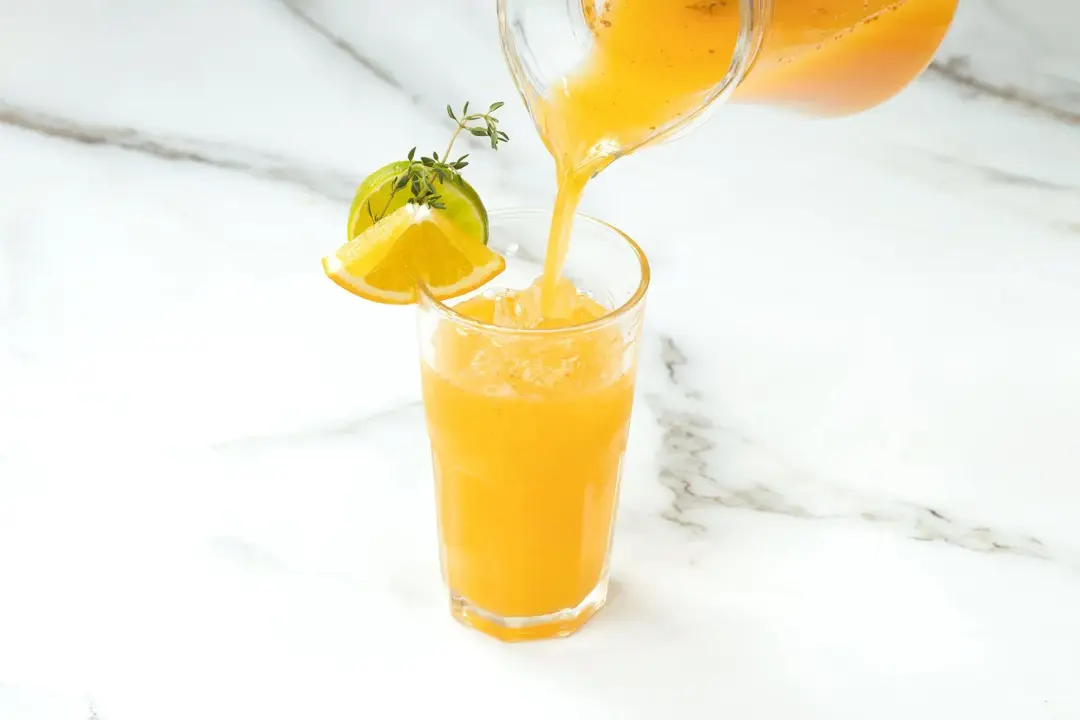 Pouring from a clear pitcher an orange cocktail into an iced glass with citrus on the rim