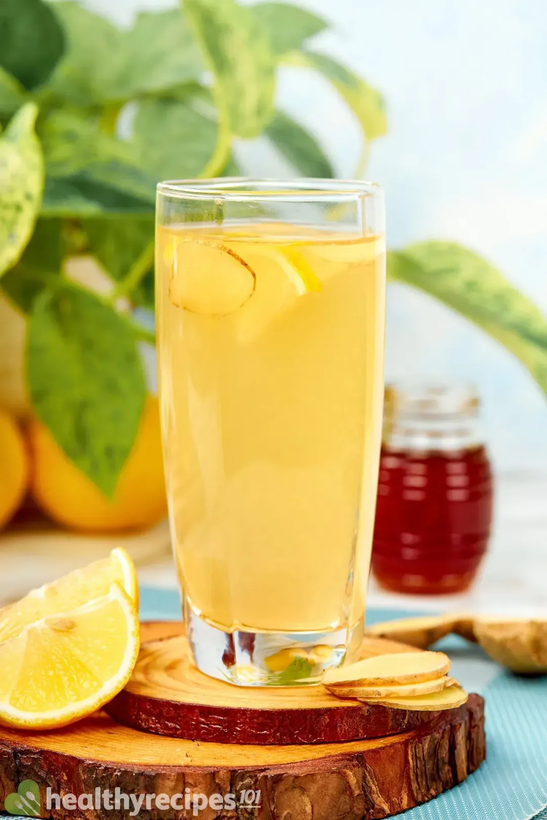 A tall glass of ginger lemon honey apple cider vinegar drink put on wooden coasters, next to some lemon wedges and ginger slices