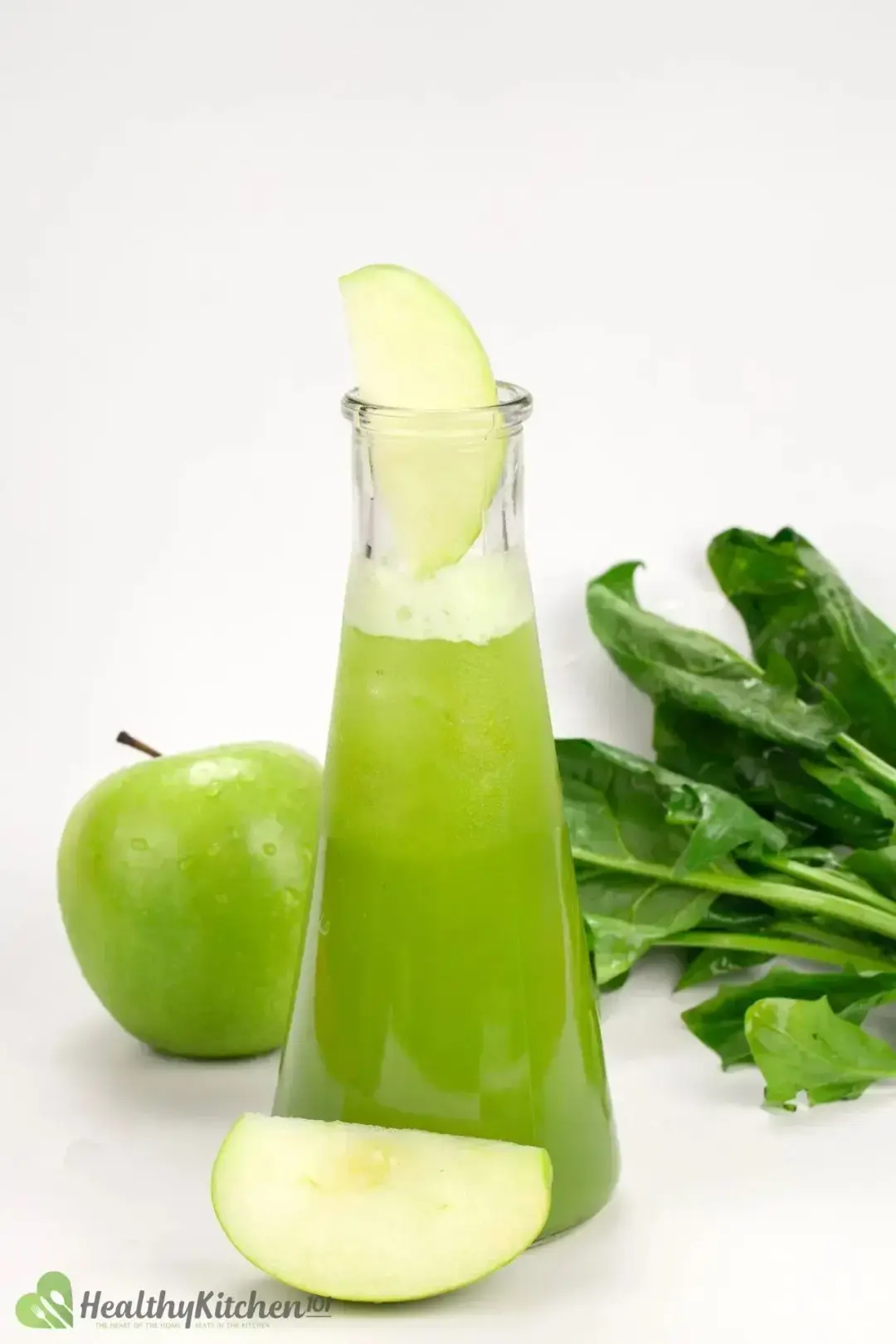A bottle-neck bottle of green apple juice with an apple wedge, green apple, and some spinach leaves around