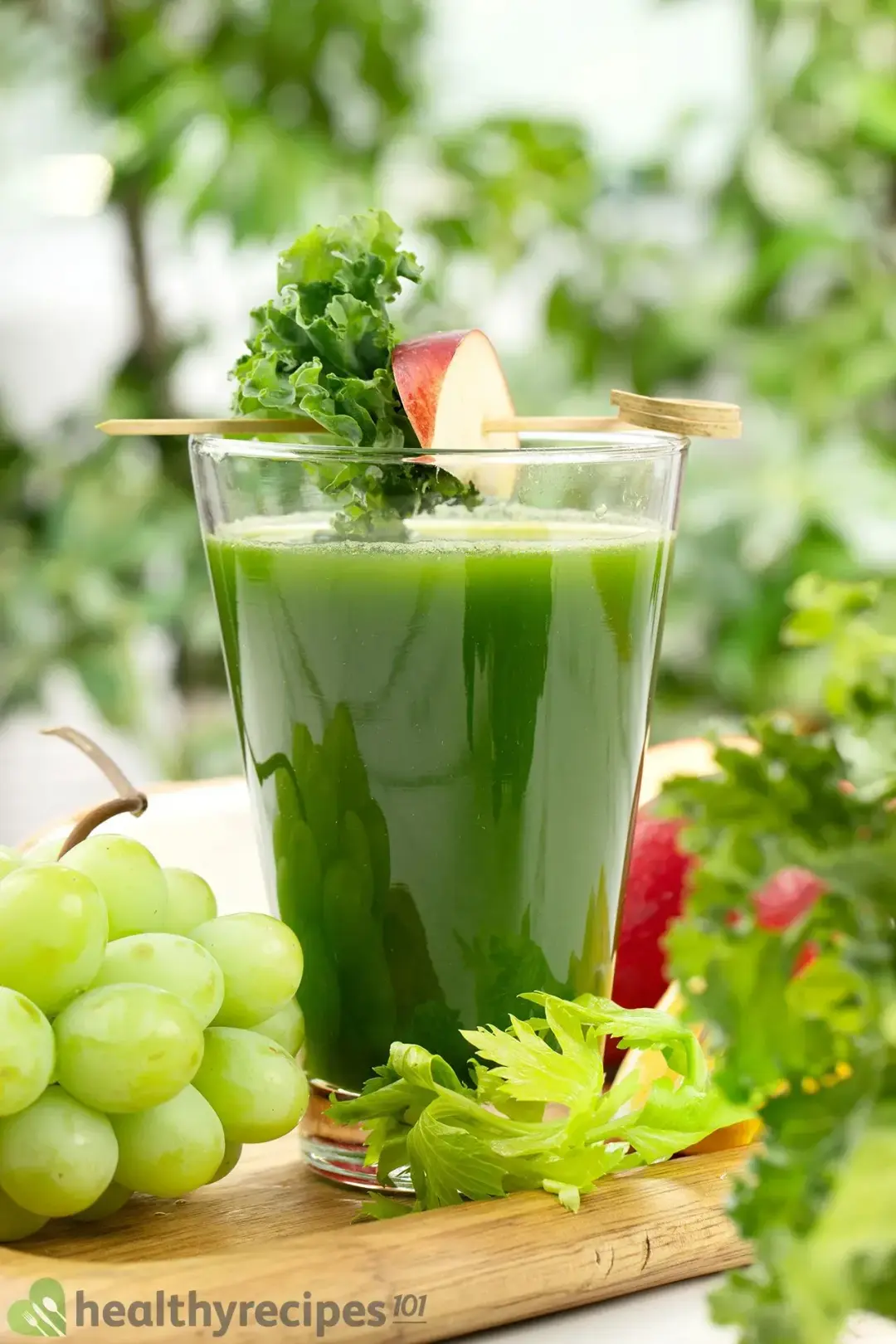 A tall glass of green machine juice on a wooden tray along with some green grapes, celery leaves, and apples