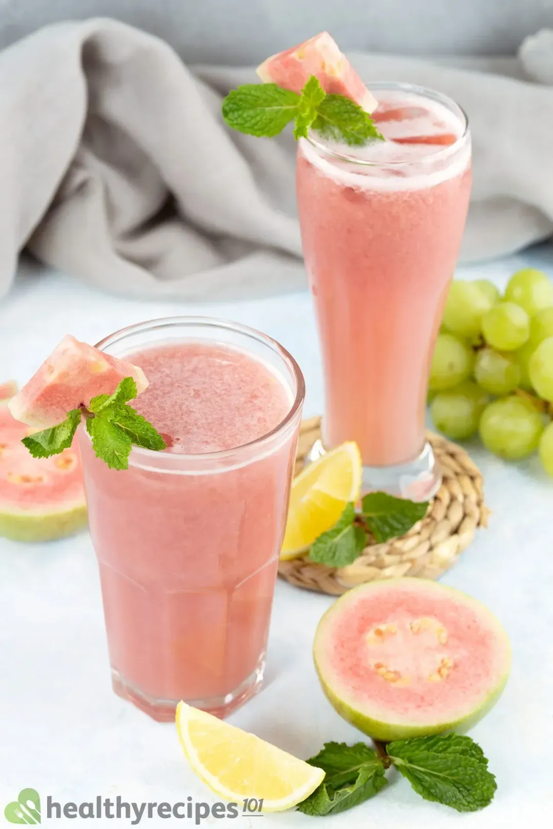 Two glasses of guava juice surrounded by pieces of guava, green grapes, and lemon slices