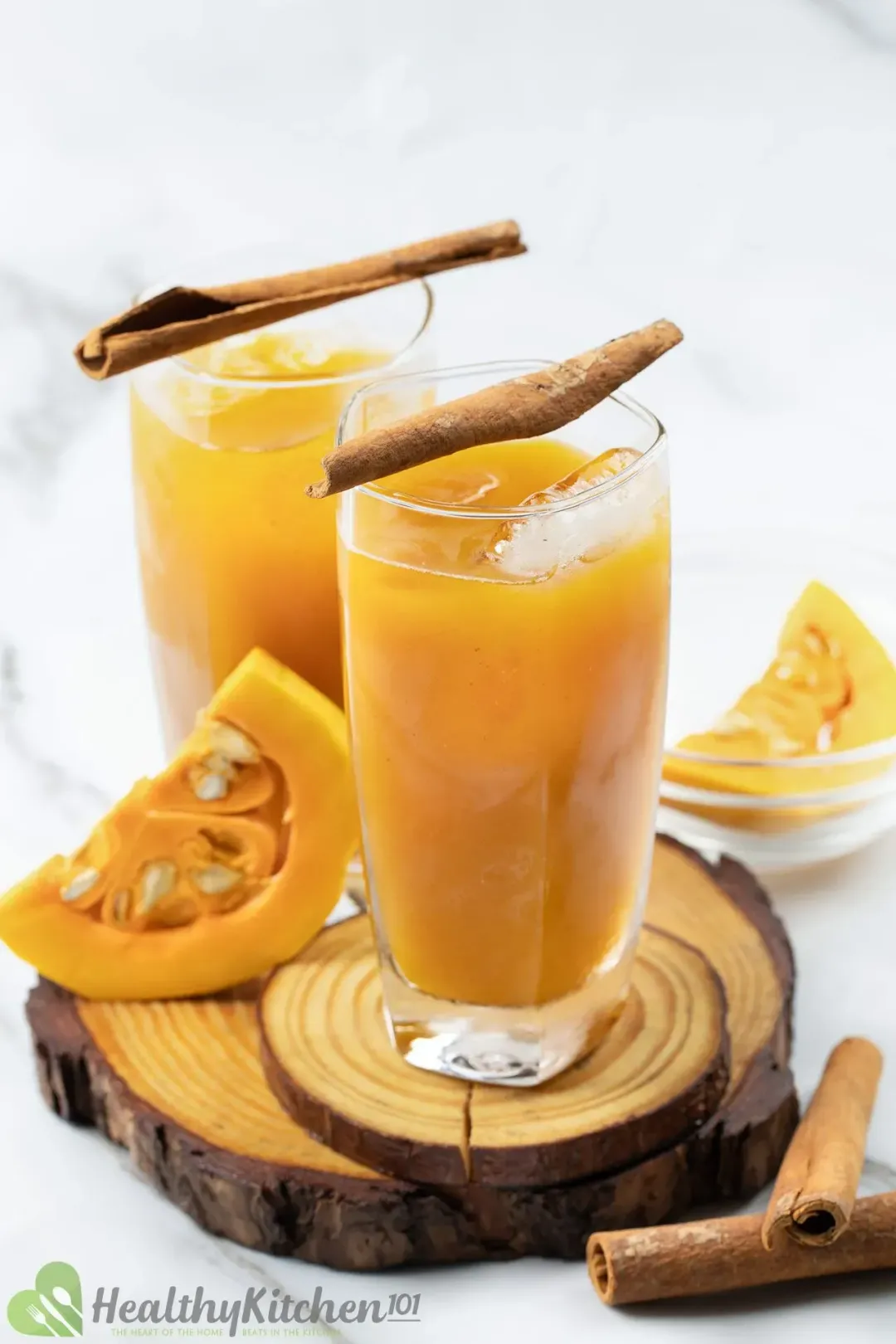 Two glasses of iced pumpkin juice decorated with cinnamon sticks on top of wooden coasters next to pumpkin slices and cinnamon sticks