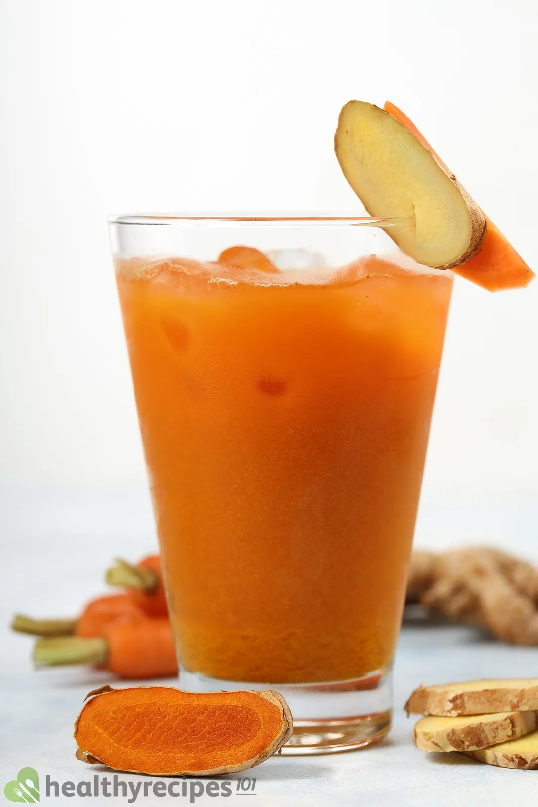 A glass of Carrot Ginger Turmeric Juice surrounded by slices of carrots and ginger.