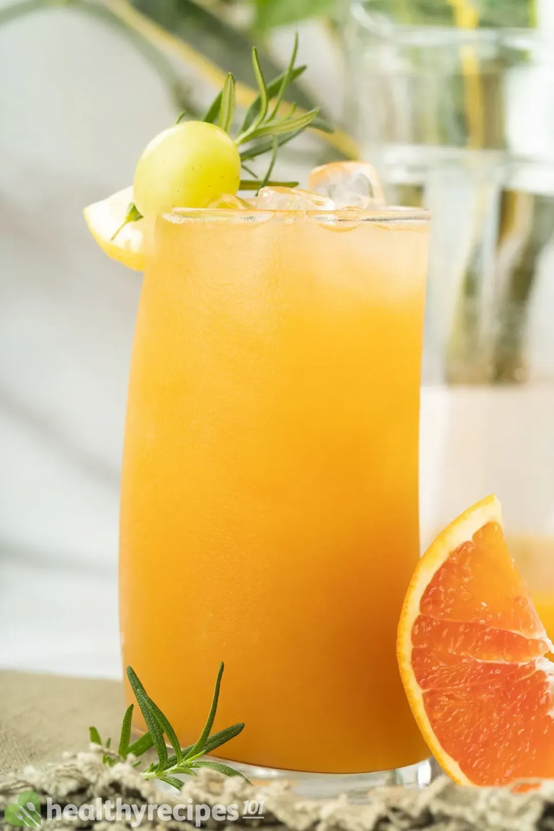 A tall glass of grapefruit juice with ice, topped with a grape, rosemary sprig, and put next to a grapefruit wedge