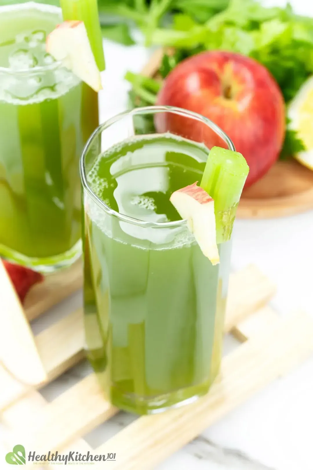 Two glasses of celery juice in tall glasses with apple slice and celery stalk as garnish, in front of a red apple