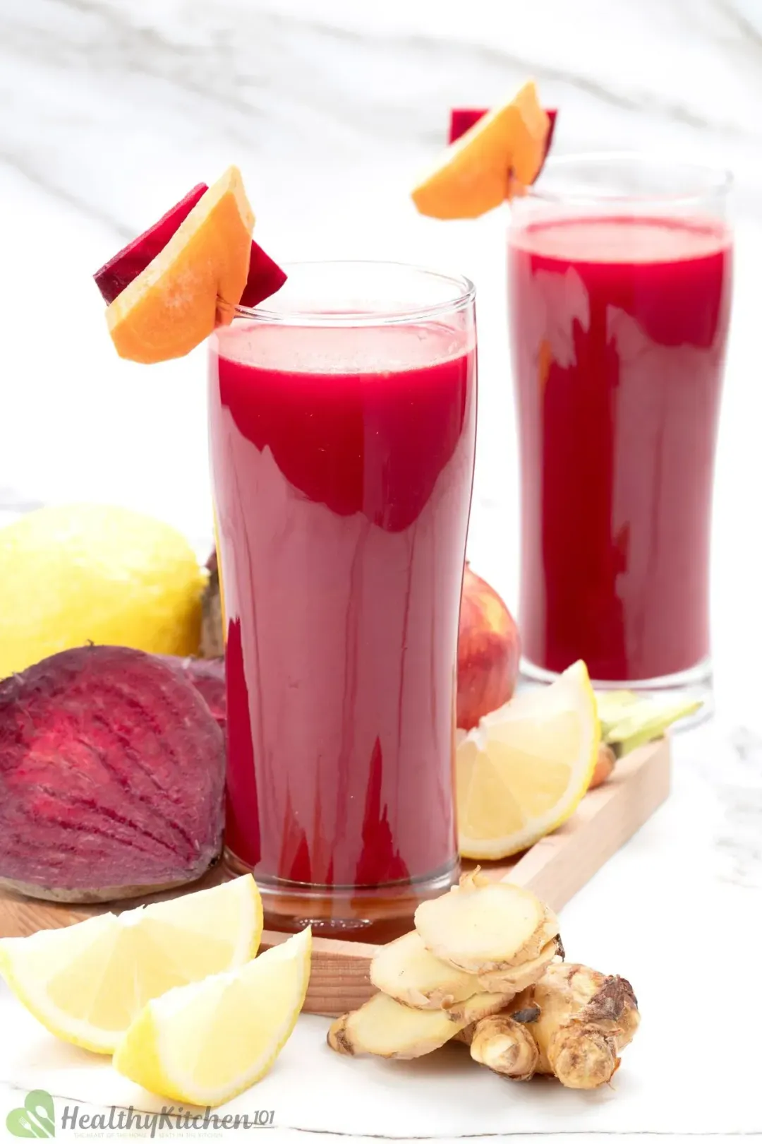 Two tall glasses of beetroot drink garnished with carrot slices, beetroot half, lemon wedges, and ginger knobs