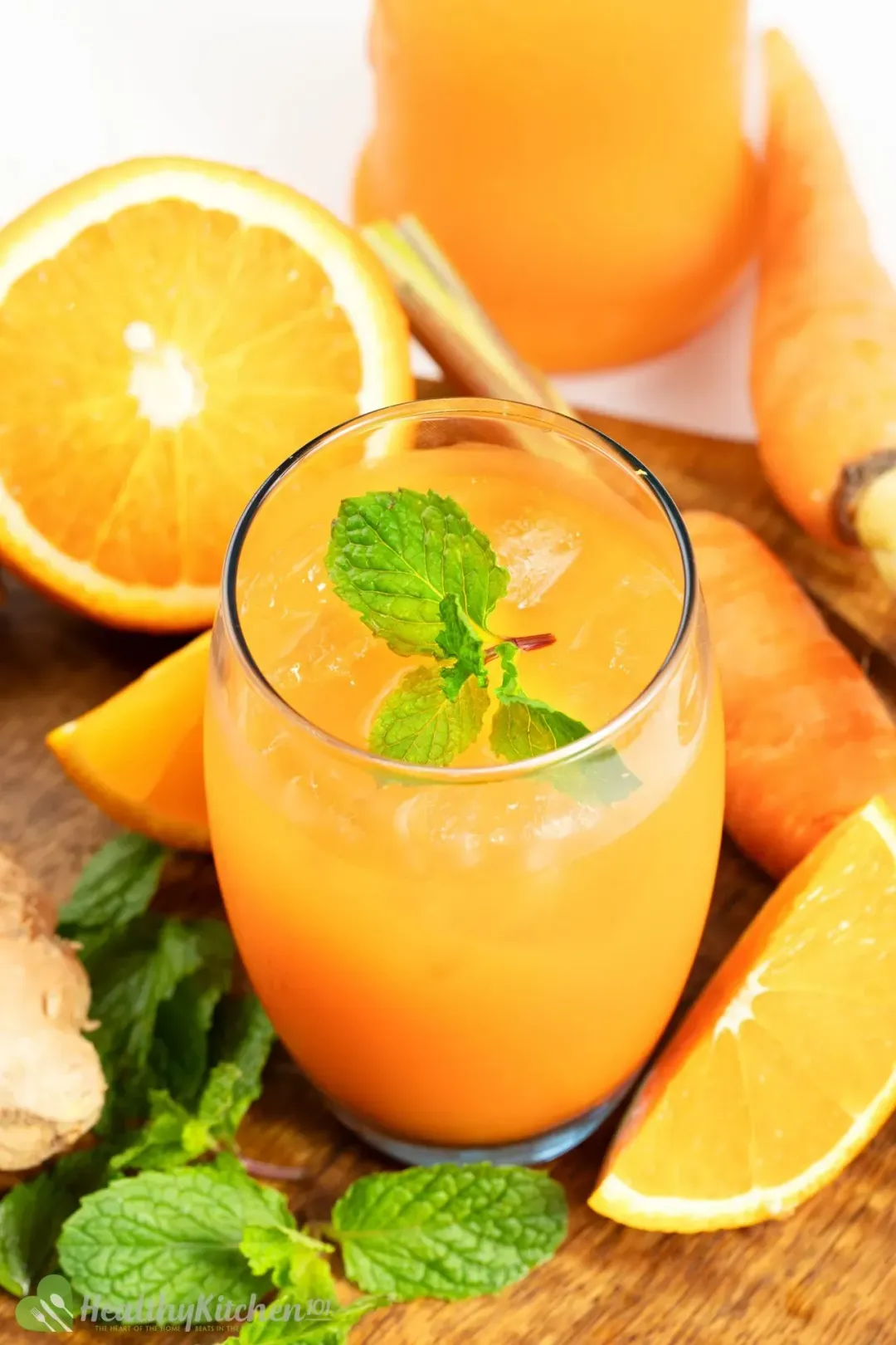 A short glass of orange carrot drink garnished with mints, orange wedges, carrots, on a wooden cutting board