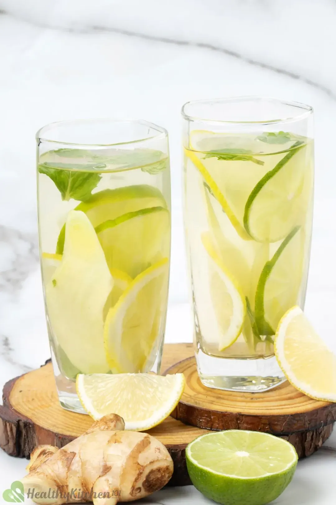 Two tall glasses filled with ginger water with sliced ginger, lemons, and limes inside, next to citrus wedges