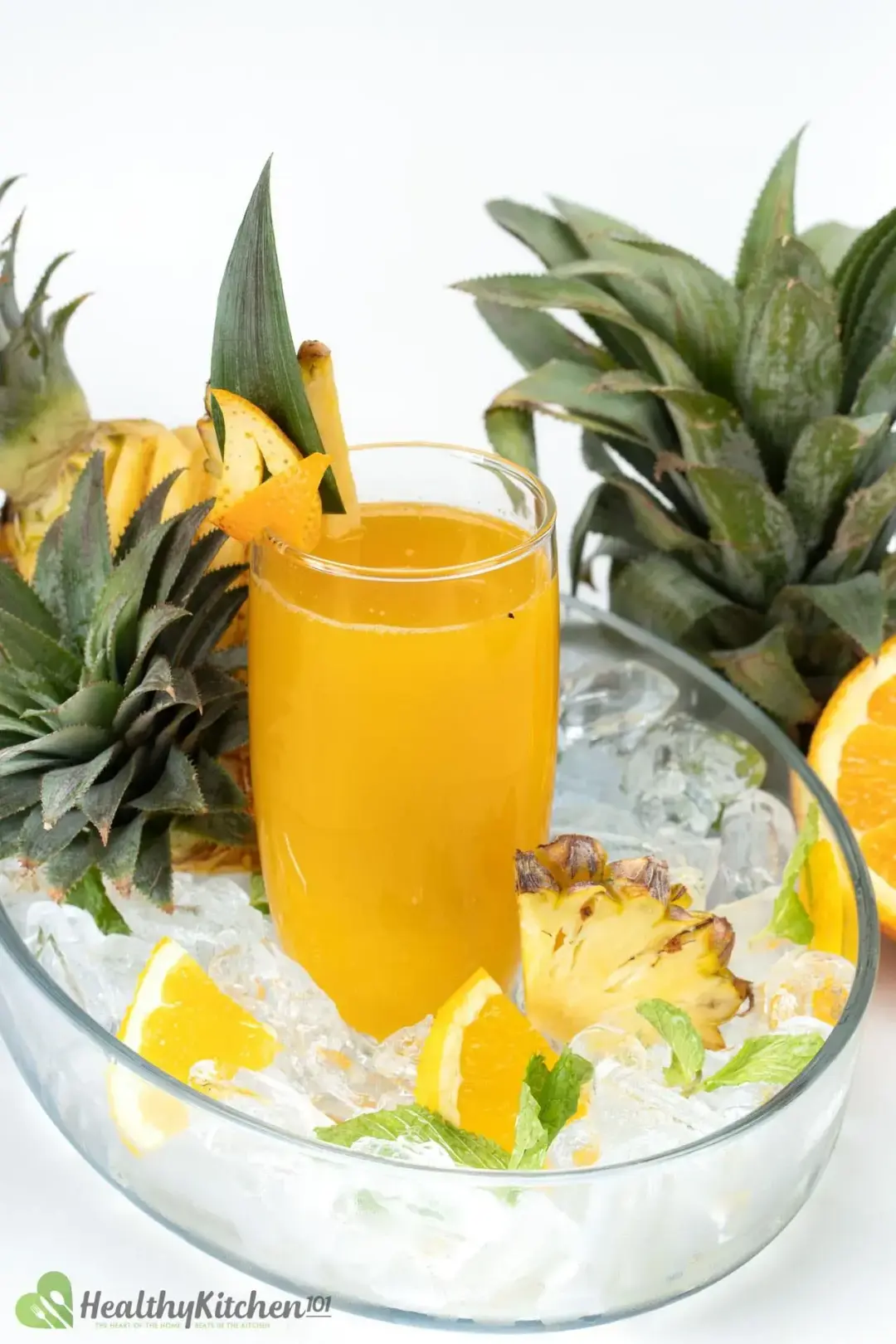 A tall glass of orange pineapple drink inside an ice bowl with orange wedges, pineapple heads, next t